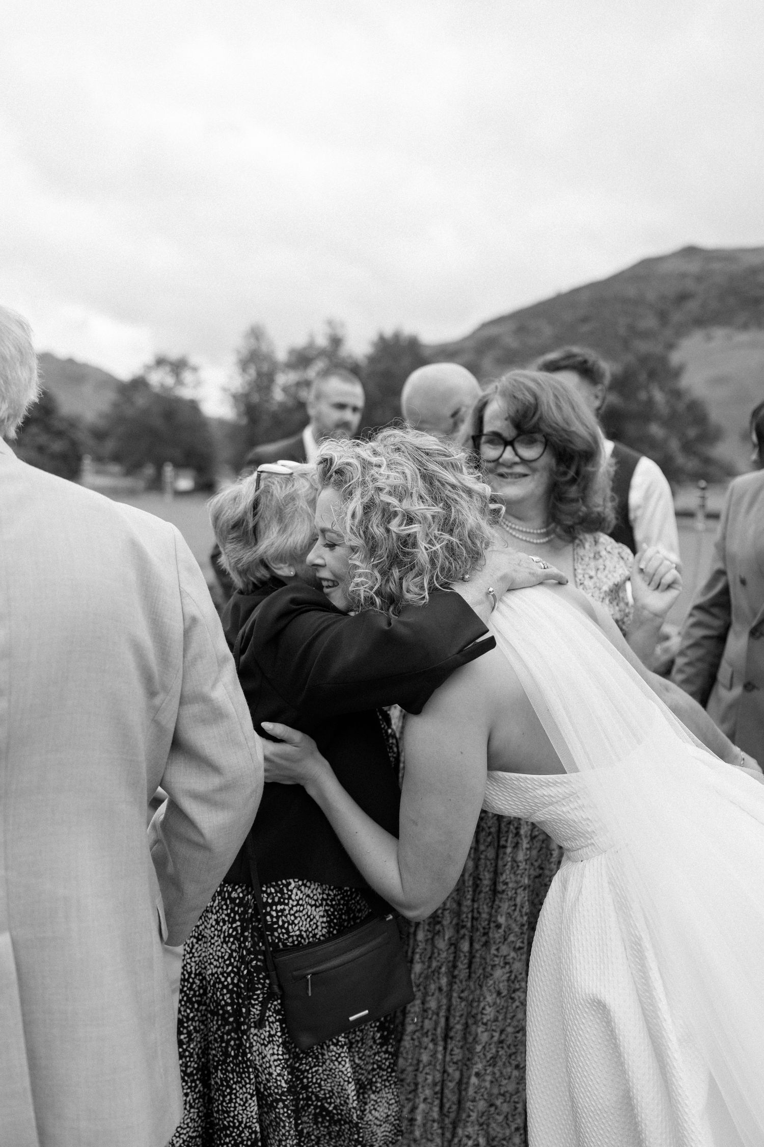 Wedding guests sharing emotional embraces during an outdoor celebration with mountains in the background.