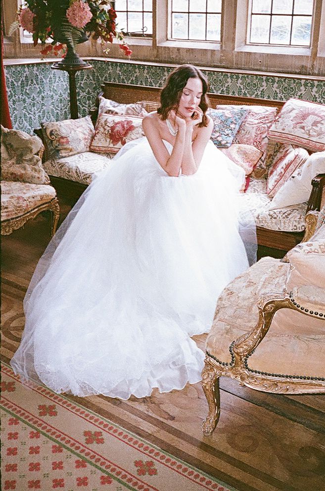 A bride in a flowing white wedding gown poses gracefully on an antique floral sofa in a vintage-style room with large windows.