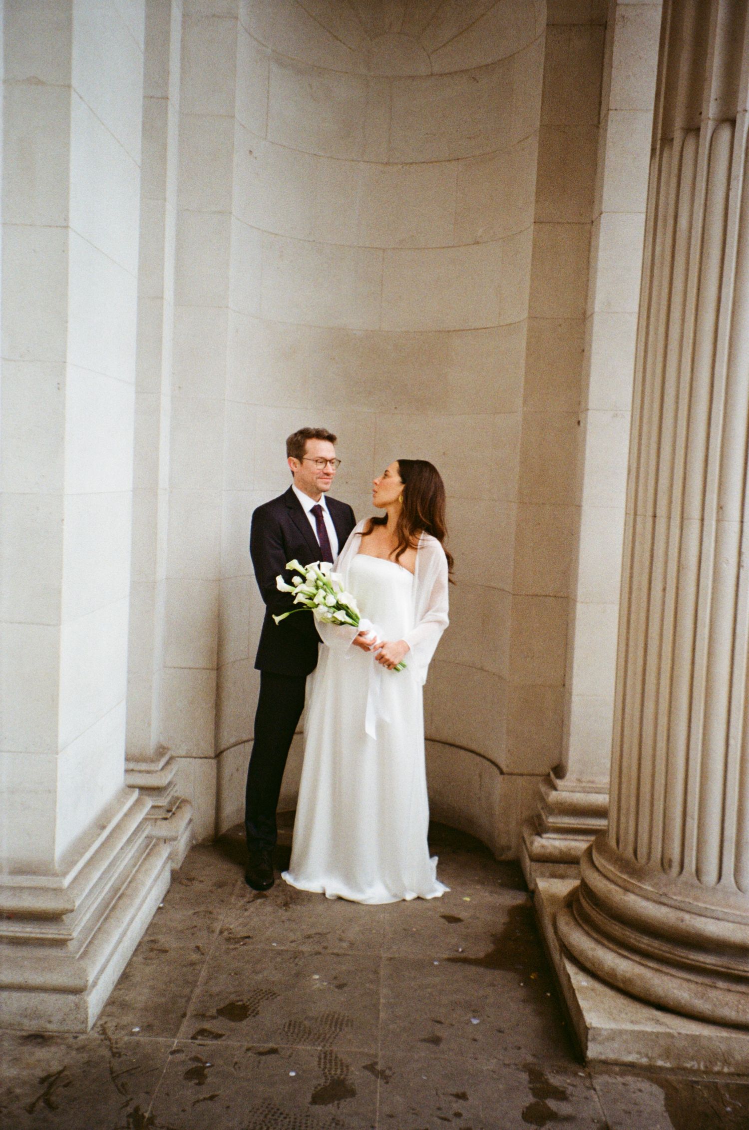 Elegant wedding couple poses between classic stone columns in a romantic architectural setting.