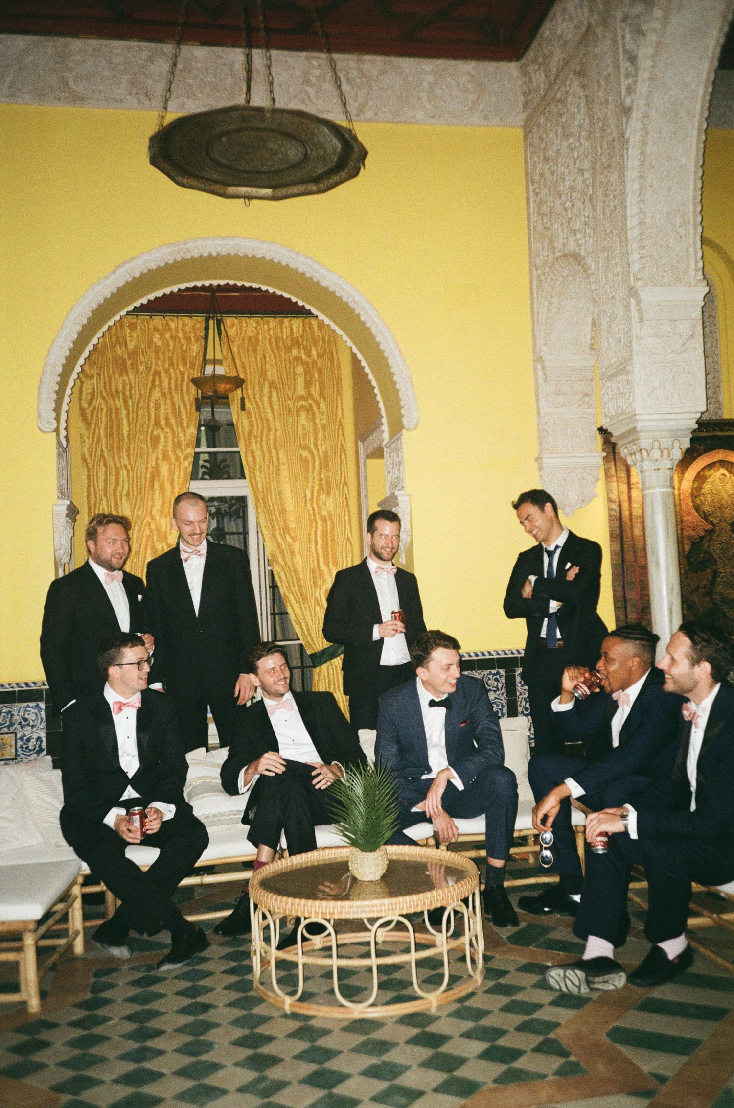 A sequence of men in black tuxedos sitting on chairs in an elegant yellow-walled room with checkered floors.