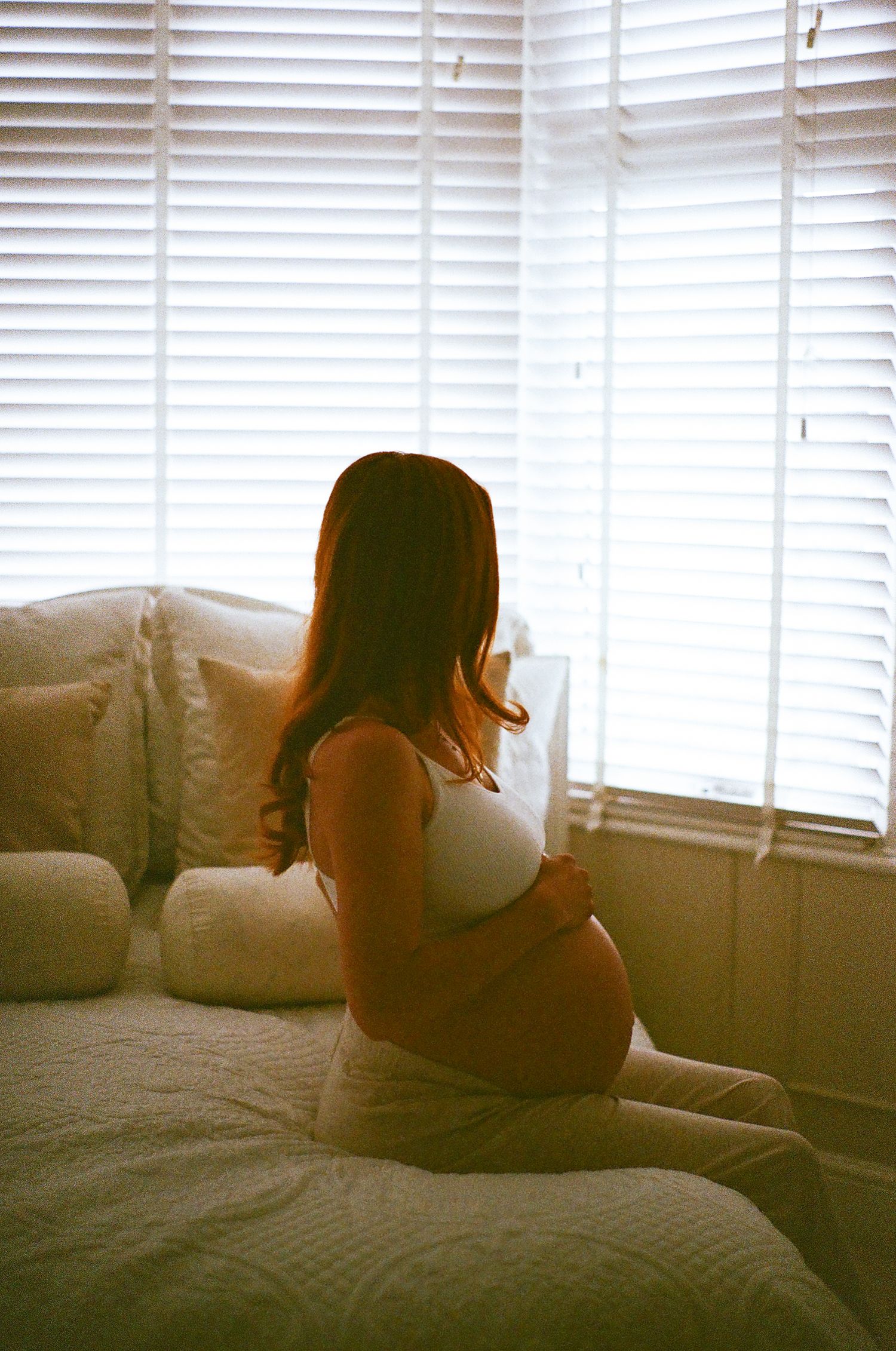 A silhouetted figure sits alone on a bed near a window with venetian blinds in a moody, atmospheric setting.