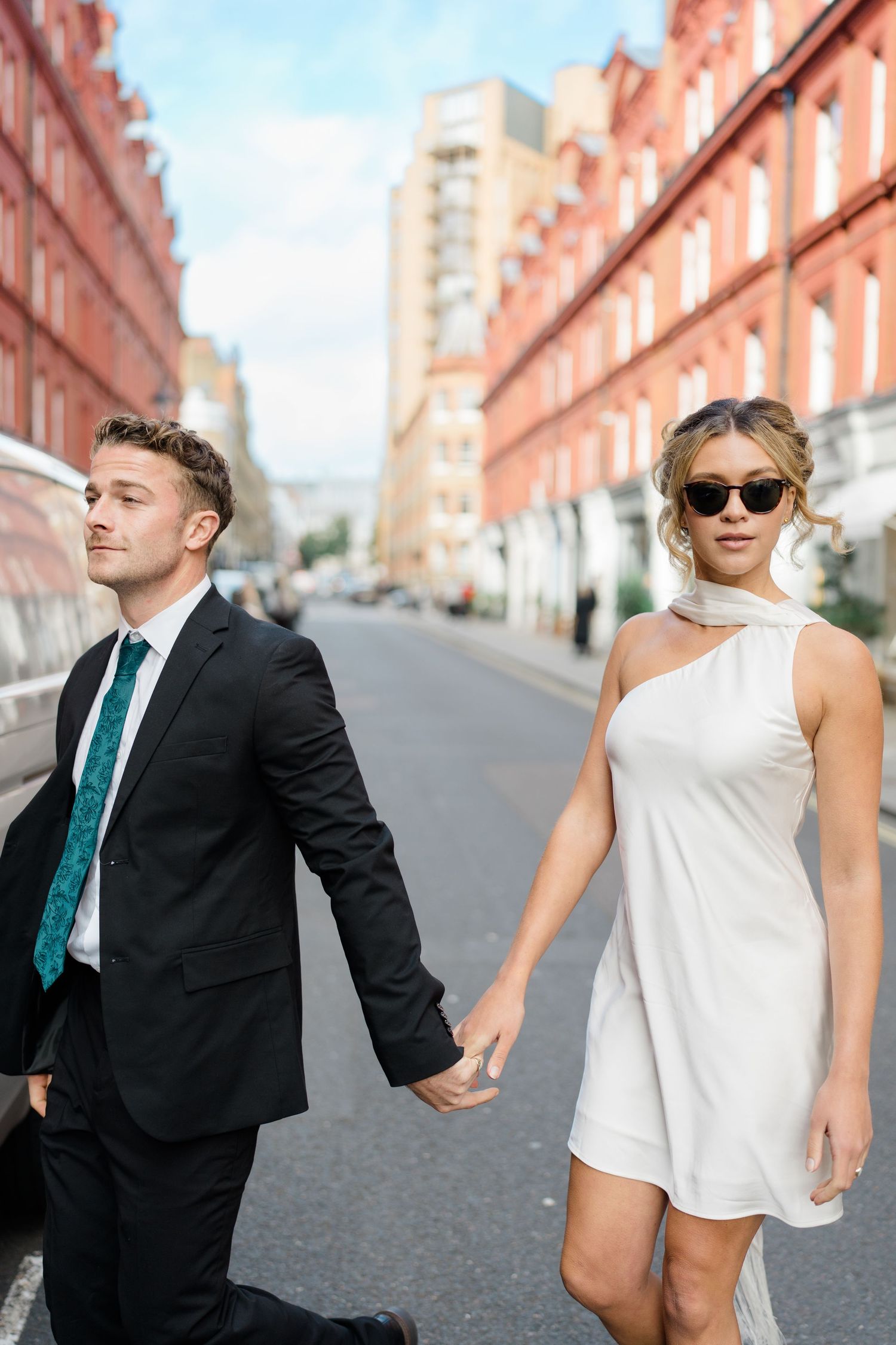 Couple in contrasting black suit and white dress pose playfully on city street.