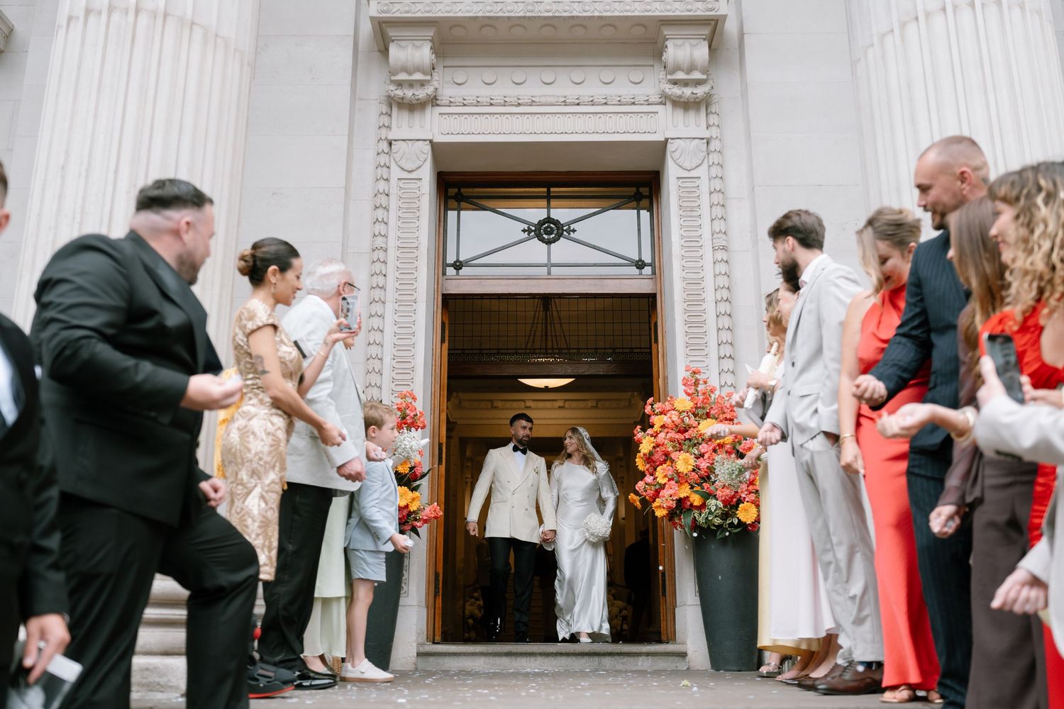 Wedding guests and party members gathered outside a classical stone building entrance.