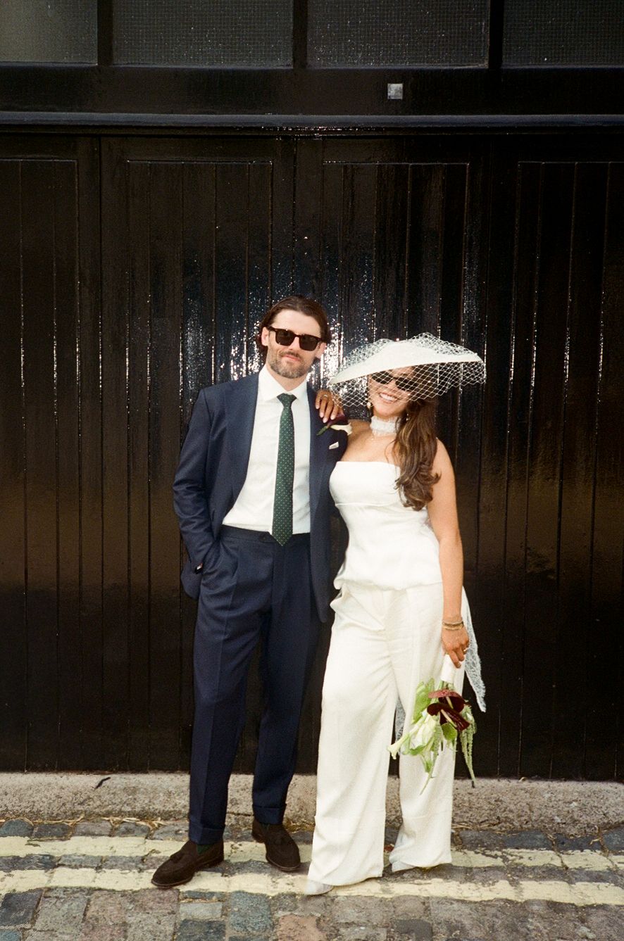 Stylish bride in white jumpsuit and veil hat with groom in navy suit posing on cobblestone street.