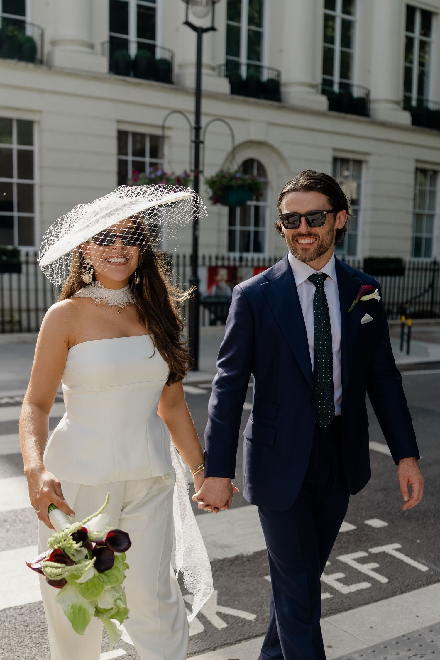 Smiling bride in white strapless suit and veil hat holds bouquet while crossing London street with groom.