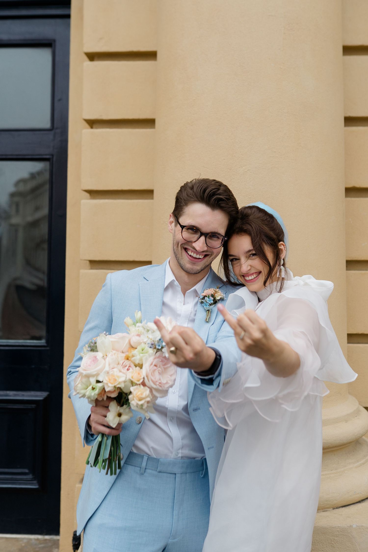 Happy newlyweds in light blue attire showing wedding rings, groom holding bouquet of pink and white roses outdoors.