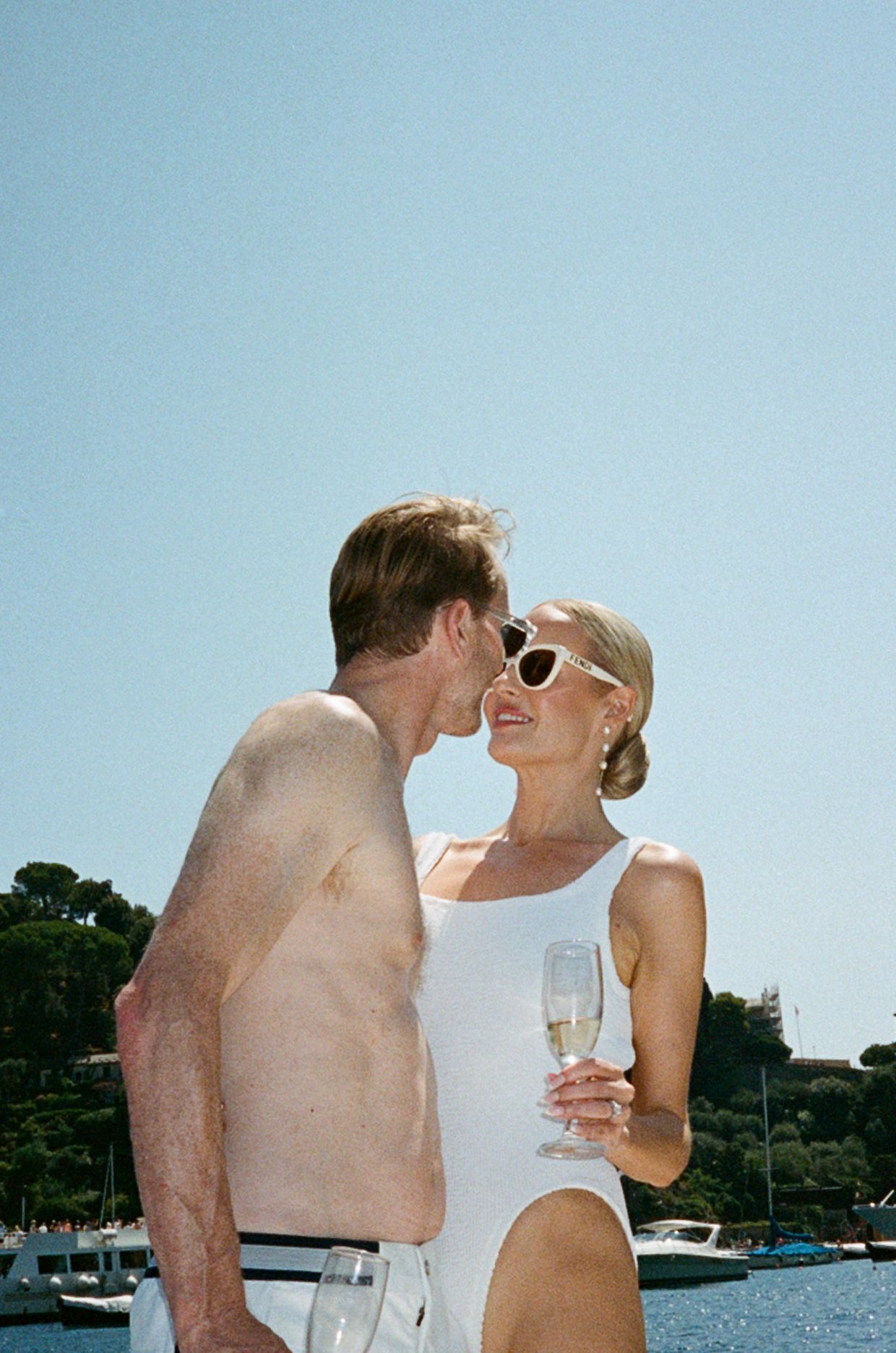 Couple nearly kissing on a sunny Mediterranean harbor, woman in white swimsuit holding champagne glass.