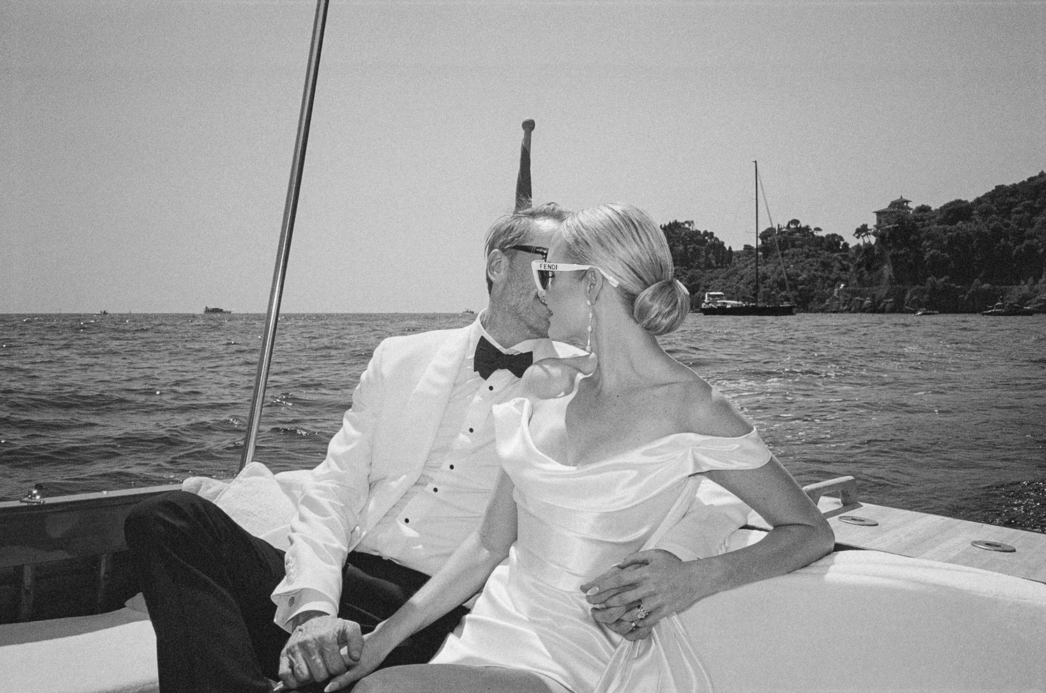 Bride and groom sharing a kiss on a boat with lush green shoreline behind them.
