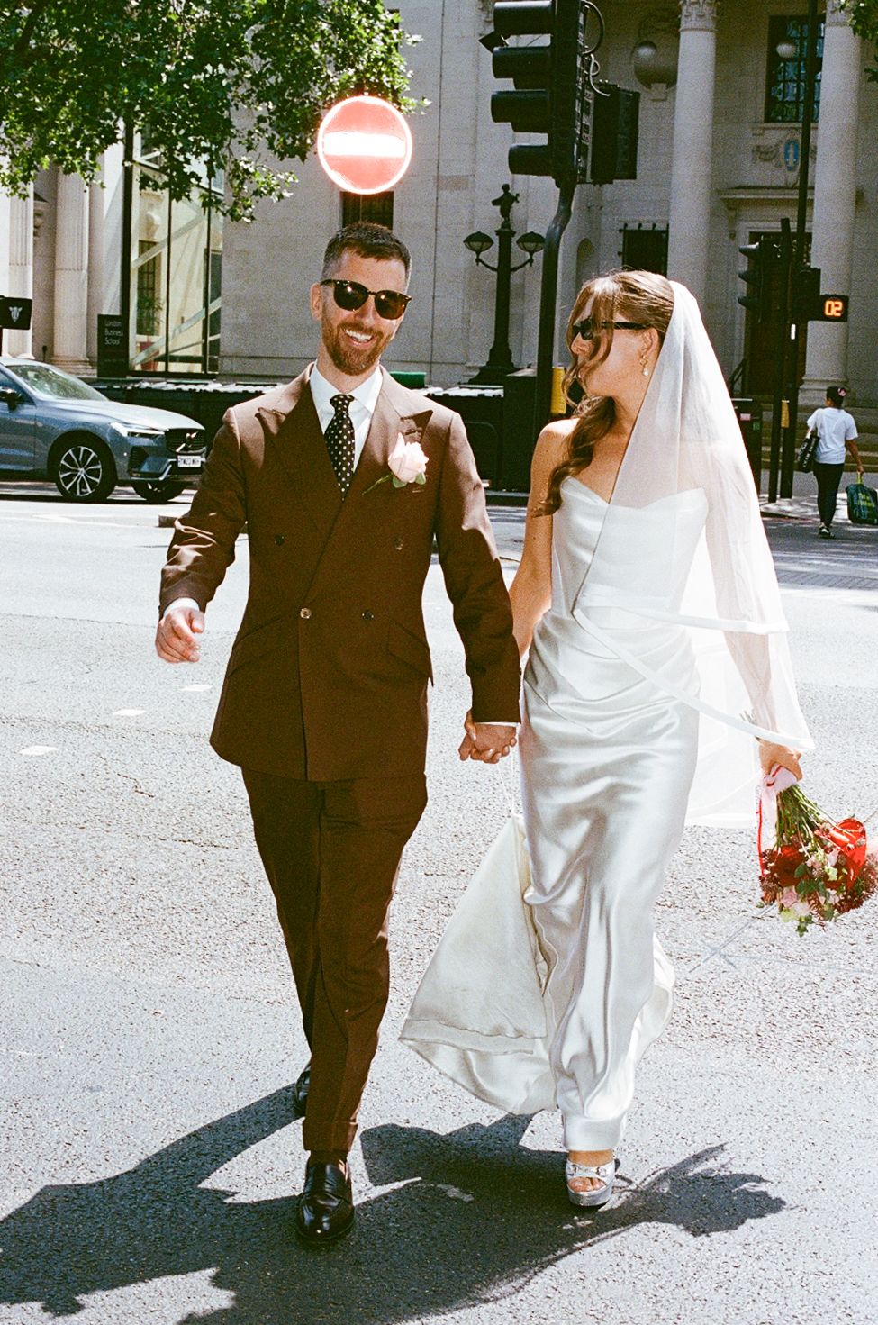Bride and groom in sunglasses walking hand in hand on sunny London street after wedding.