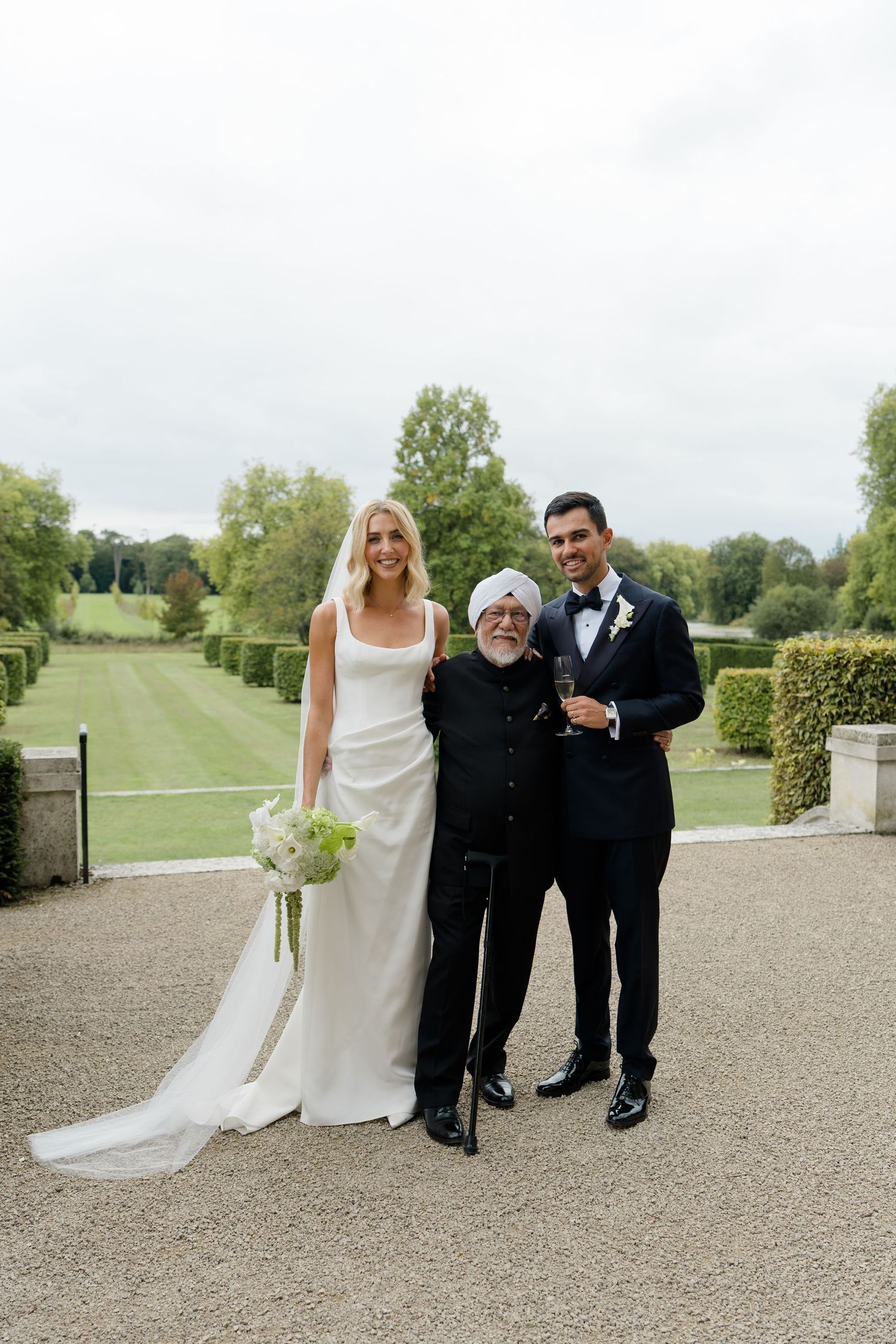 Bride in white gown and groom in navy suit pose with elderly guest in formal garden setting.