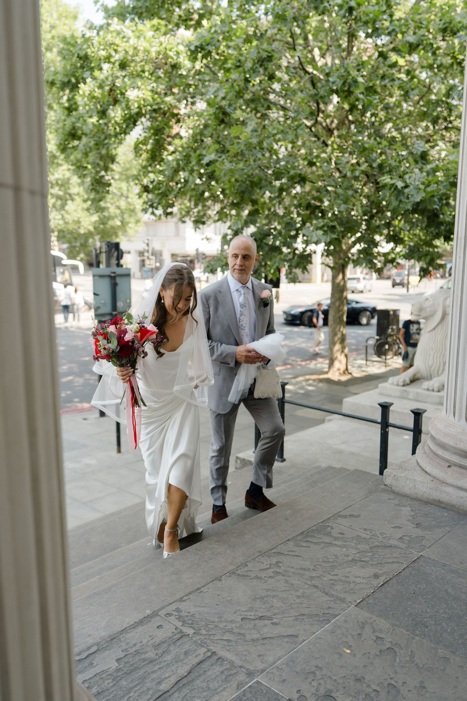 Smiling bride in white dress with red bouquet ascending stone steps with older gentleman escort.