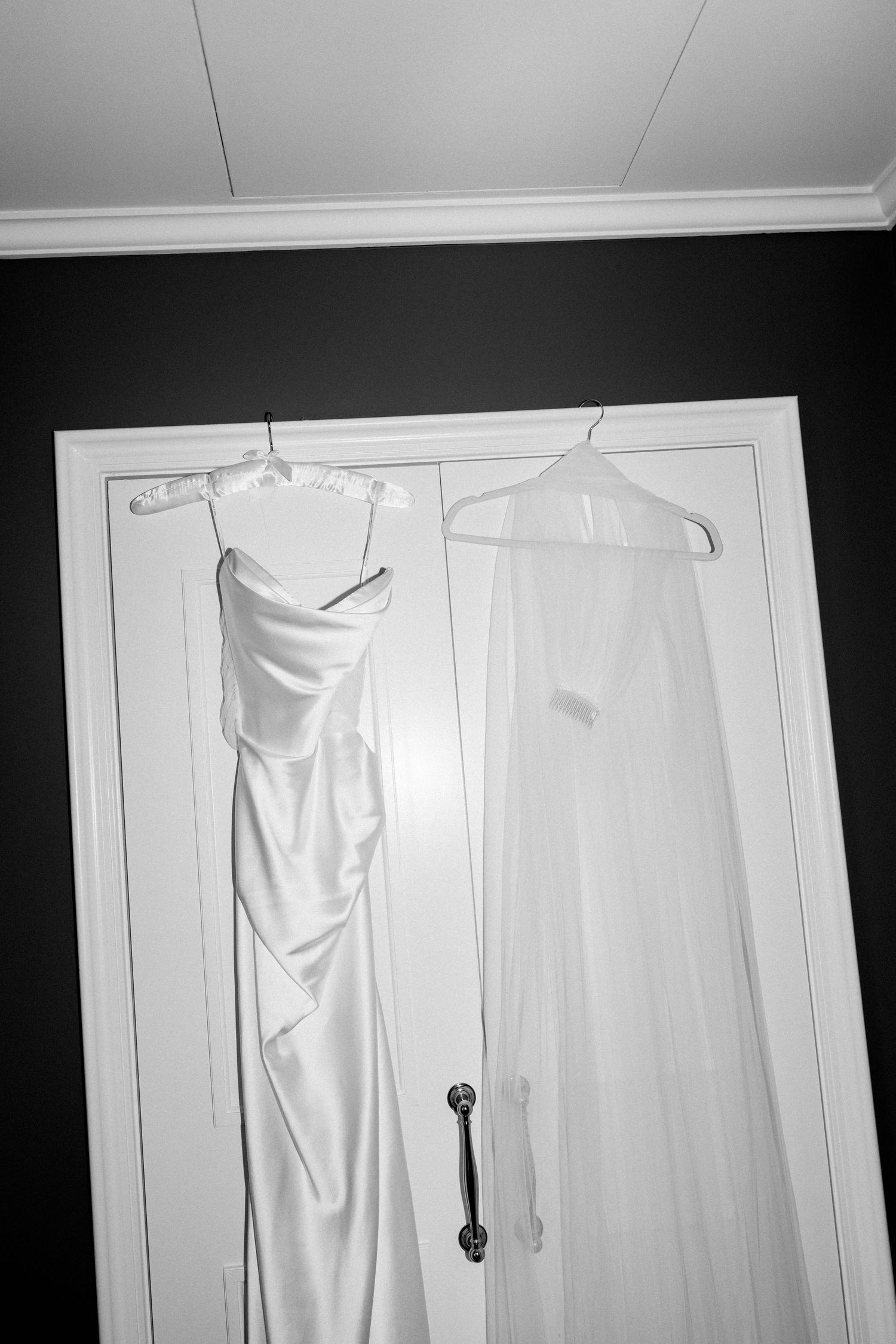 Black and white photo of a wedding dress and veil hanging on a white door before the ceremony.