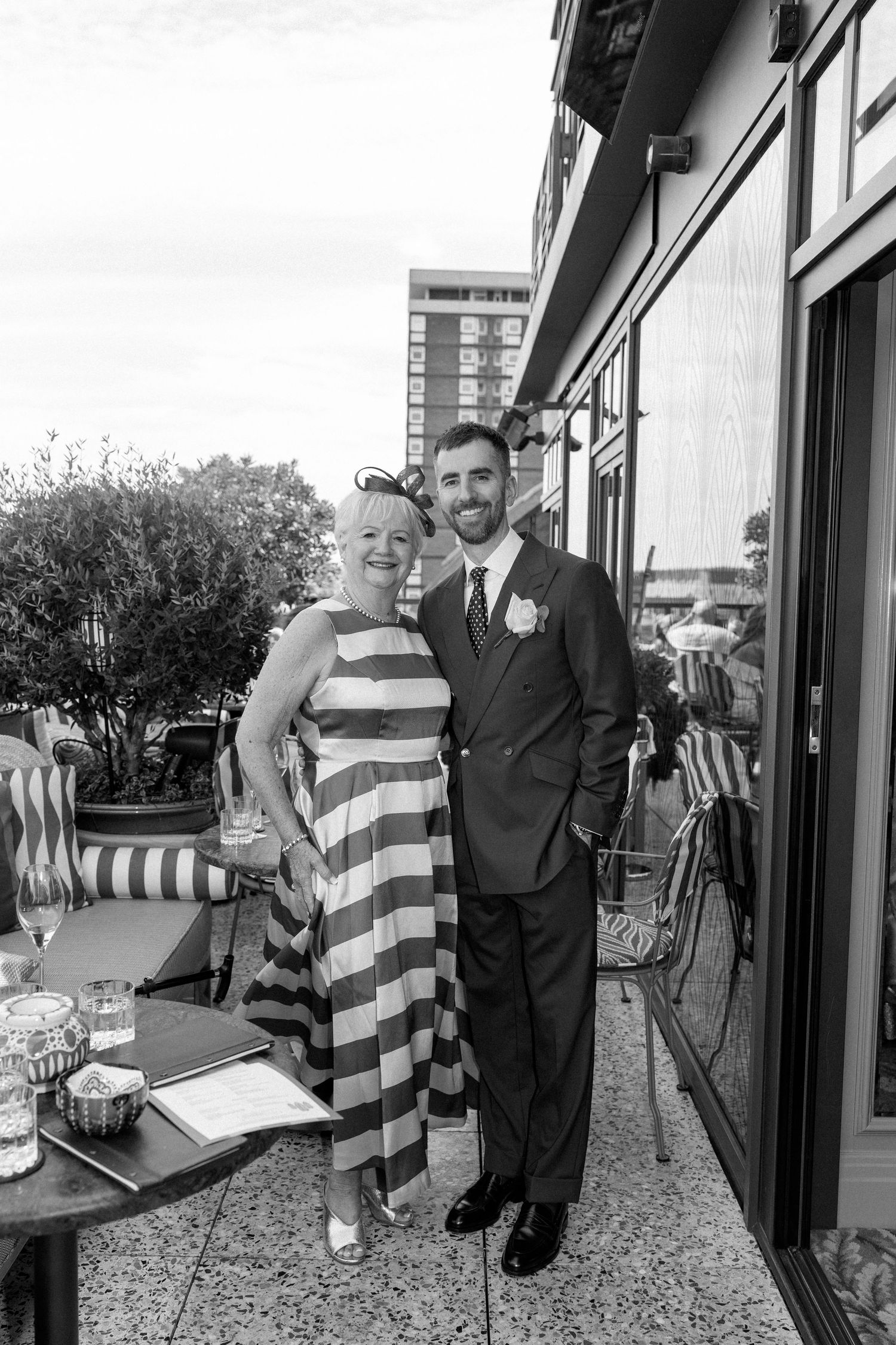 Groom in dark suit poses with smiling woman in striped dress on outdoor terrace at wedding reception.