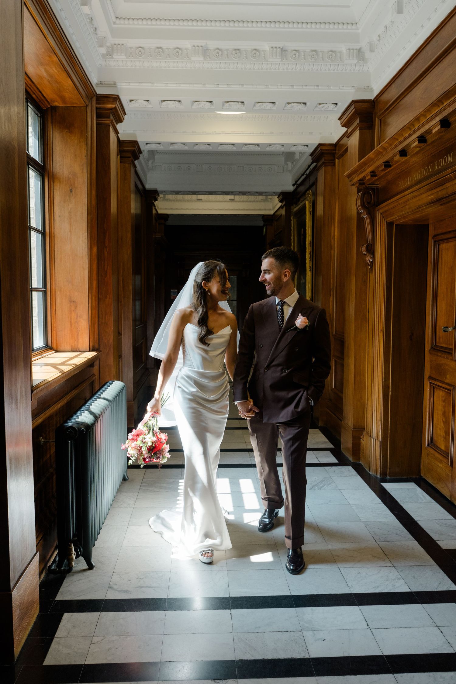 Bride and groom walk hand in hand through sunlit wood-paneled corridor after wedding ceremony.