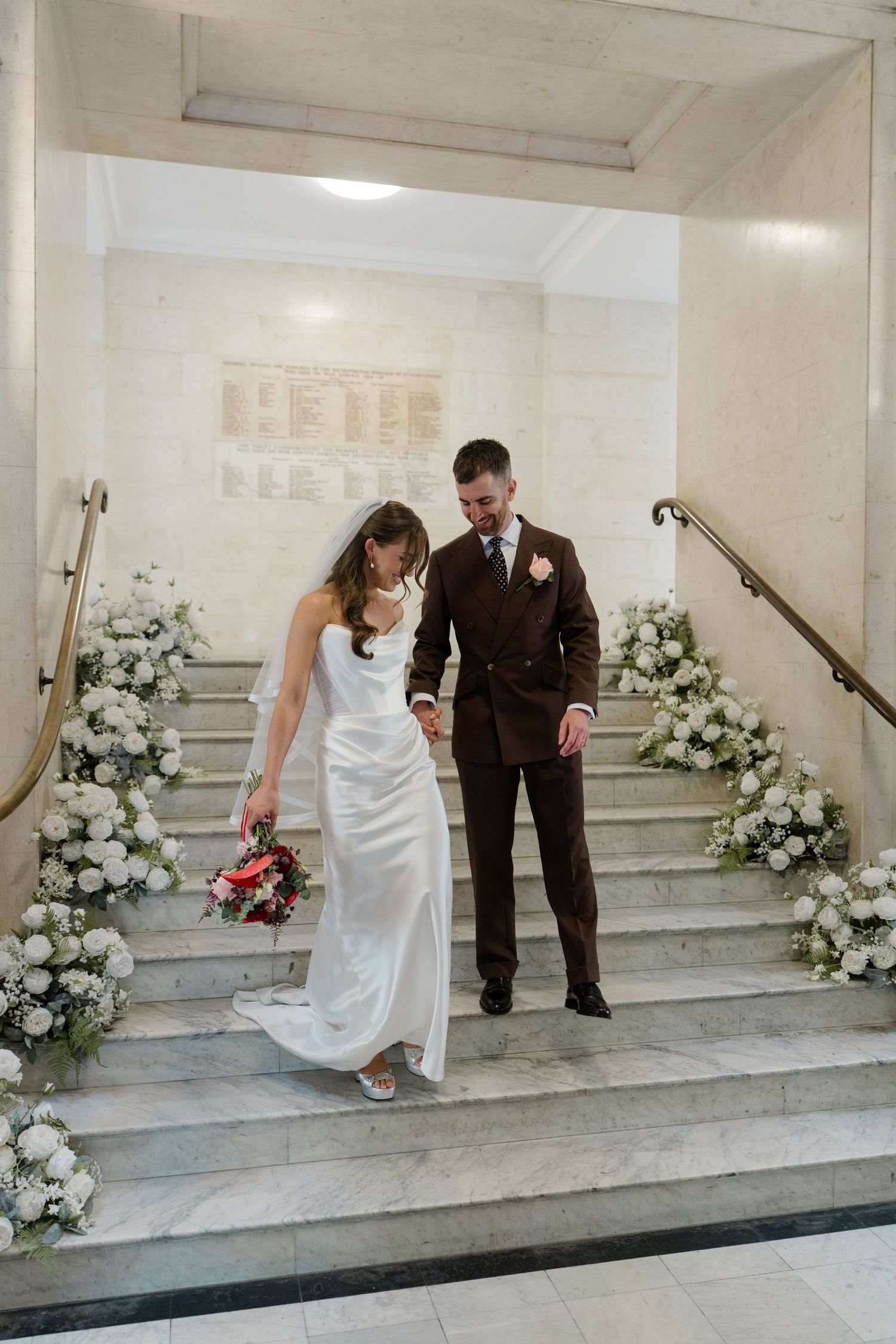 Bride and groom walking down marble stairs adorned with white floral arrangements at wedding venue.