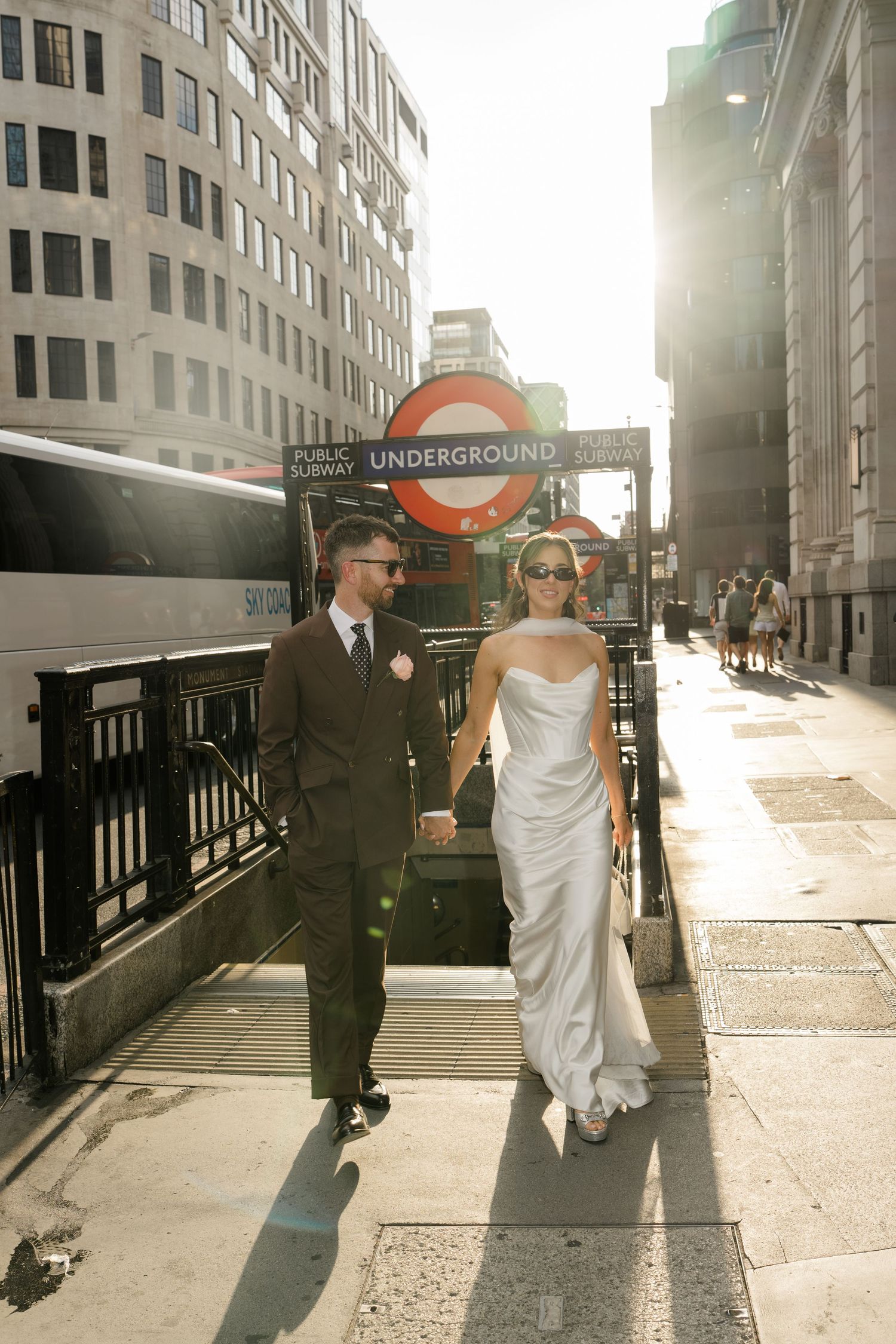 Bride and groom in sunglasses walk hand-in-hand past London Underground station entrance in golden sunlight.