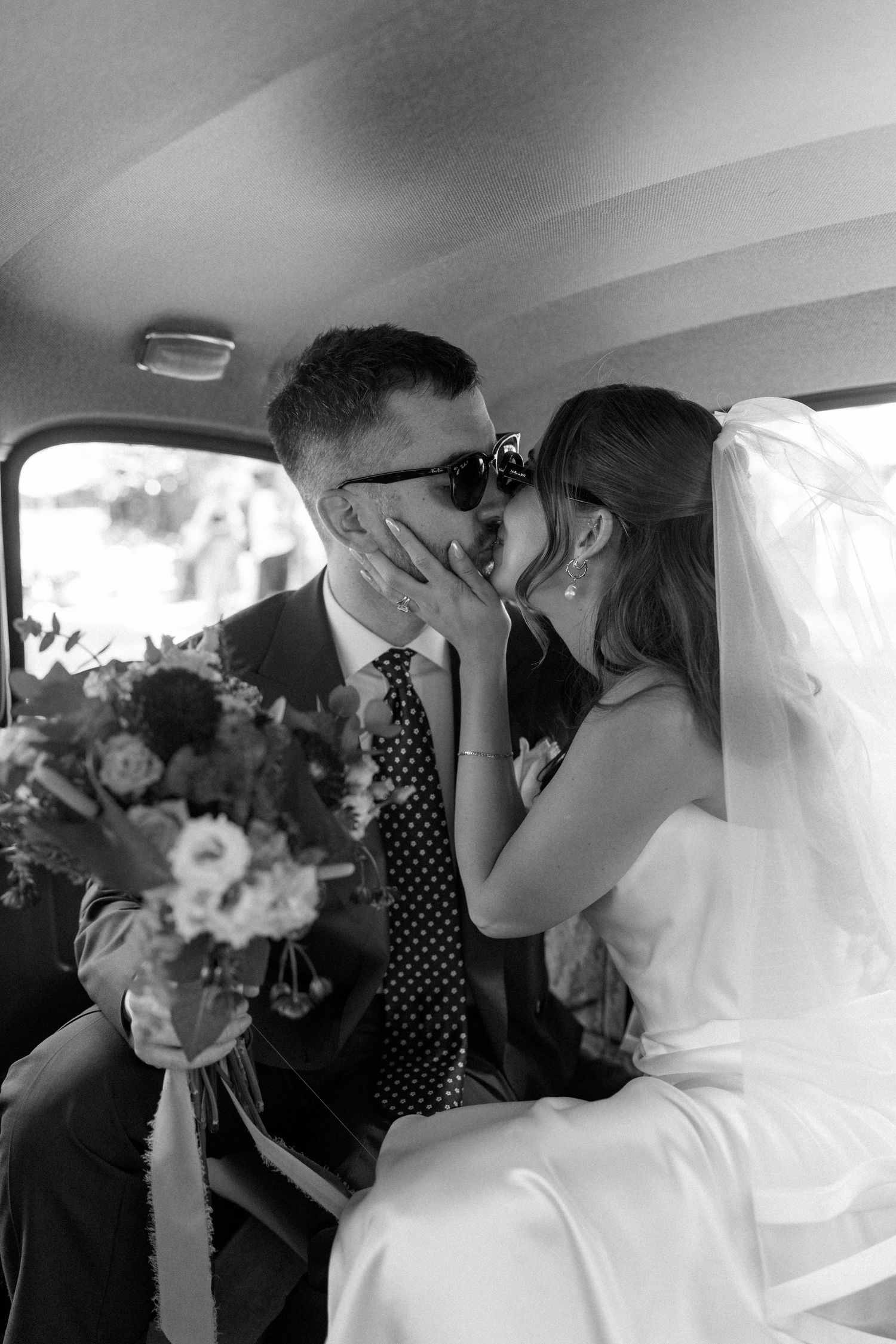 Newlyweds sharing a kiss inside a vintage car, bride in white satin dress and groom in suit with polka dot tie.