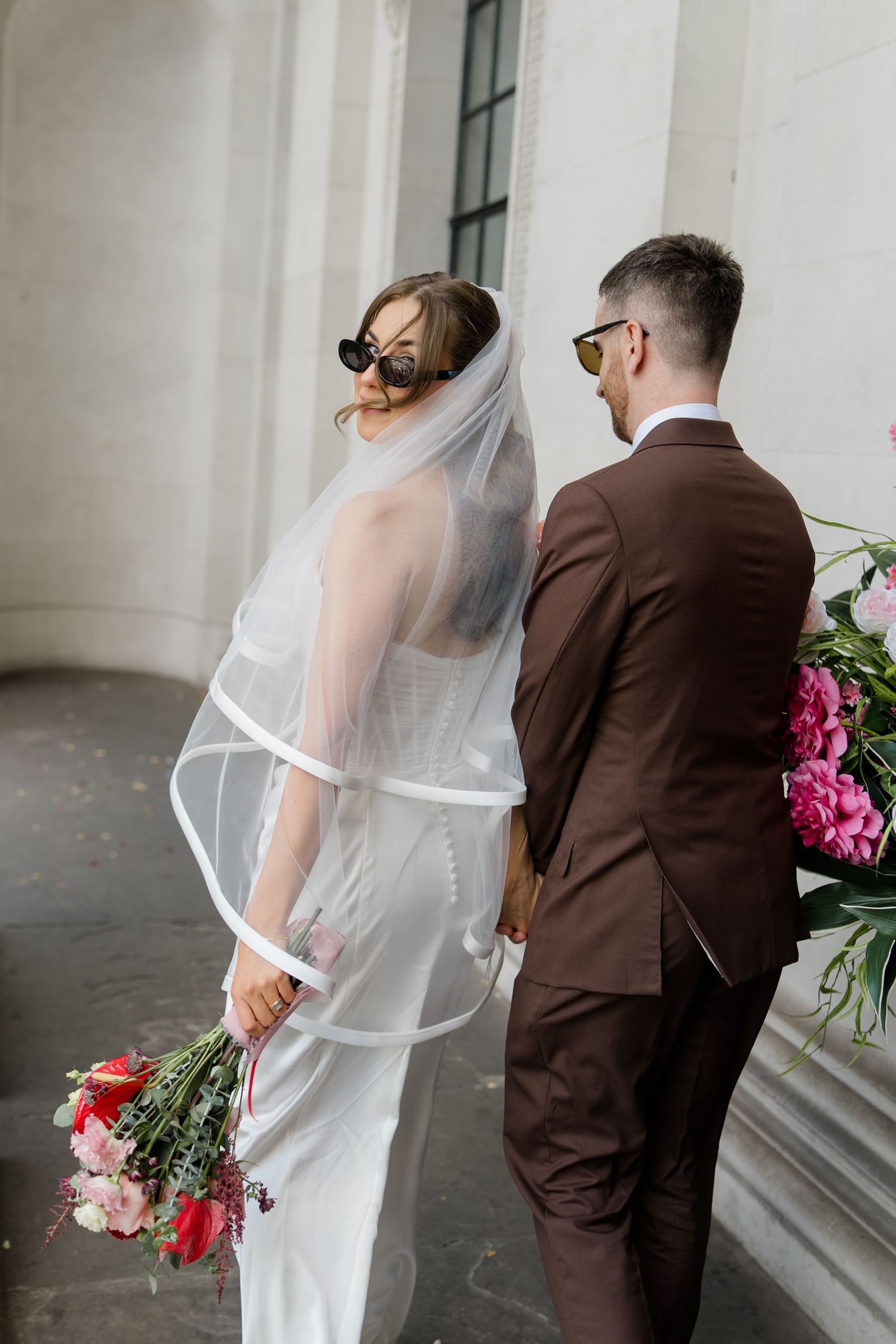 Bride in white gown and veil holding pink bouquet stands back-to-back with groom in brown suit wearing sunglasses.