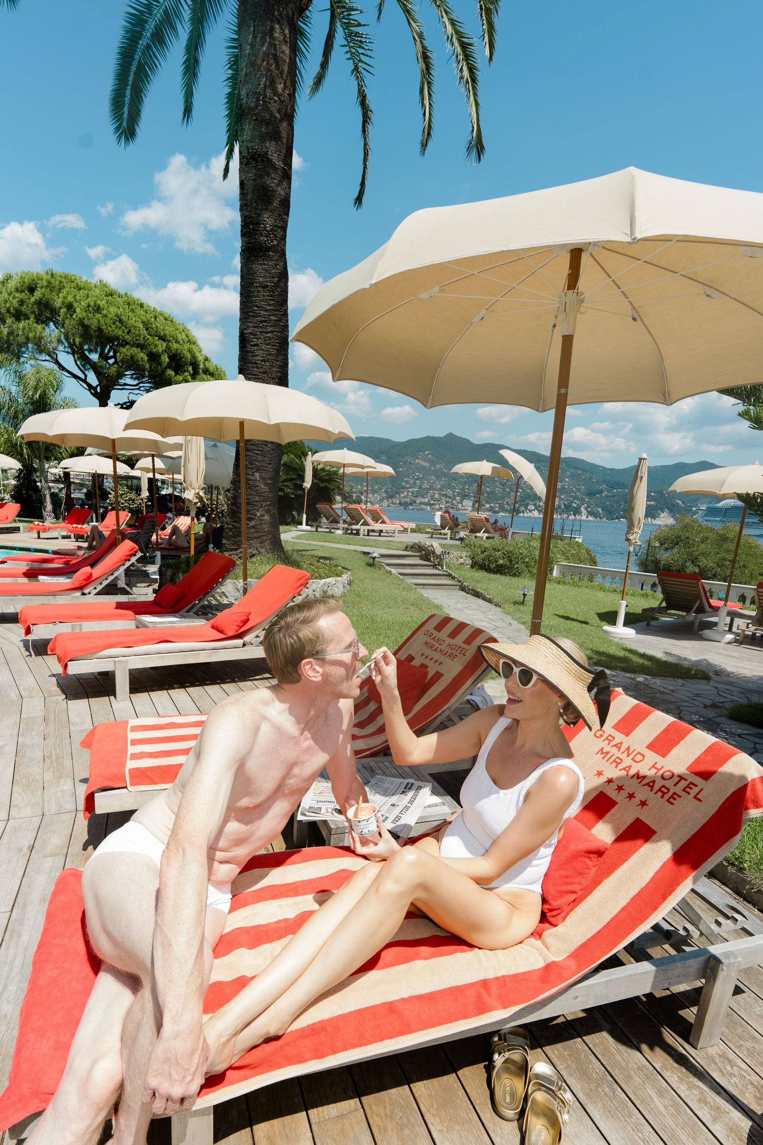 Newlywed couple relaxing on red striped sun loungers under umbrellas at a luxury Italian seaside resort.