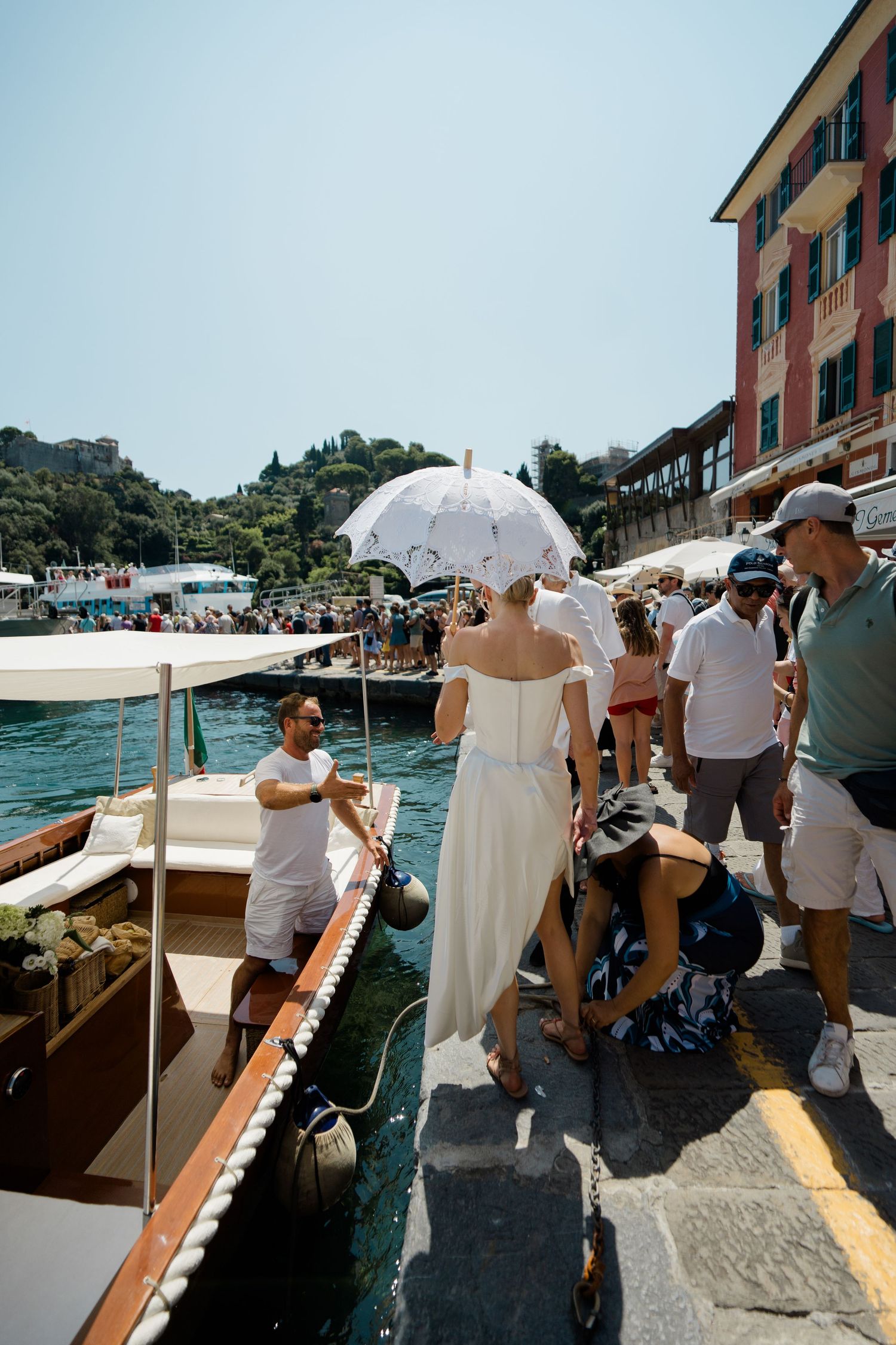 Bride in white dress holding parasol boarding a boat at the colorful Portofino harbor waterfront.
