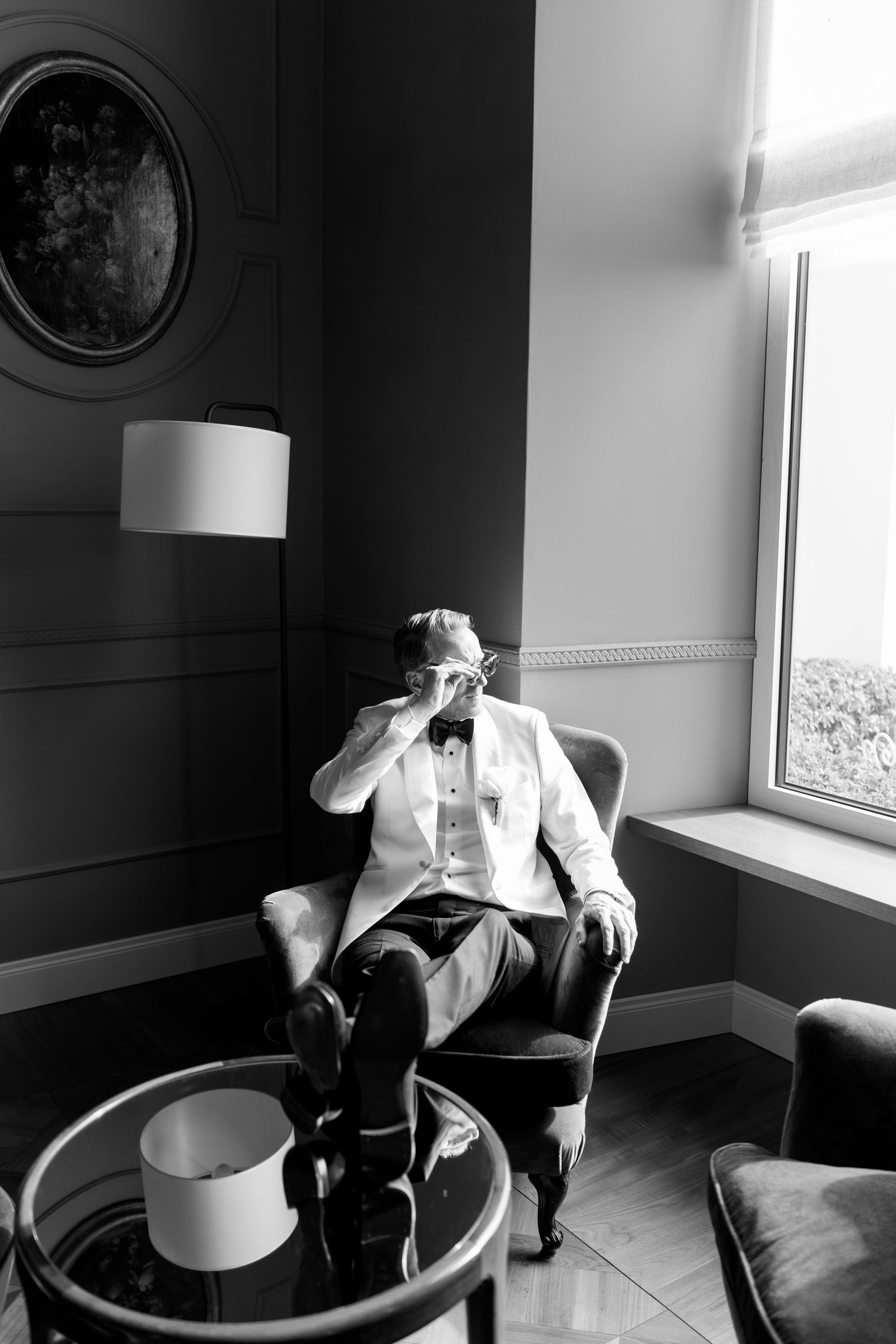 Groom in white tuxedo jacket sitting in armchair drinking coffee in elegant black and white interior.