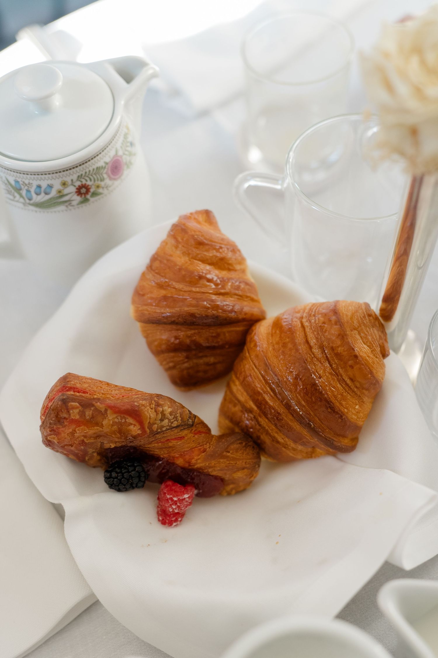 Golden croissants and a chocolate pastry with fresh berries on a white plate, served with a floral teapot for breakfast.