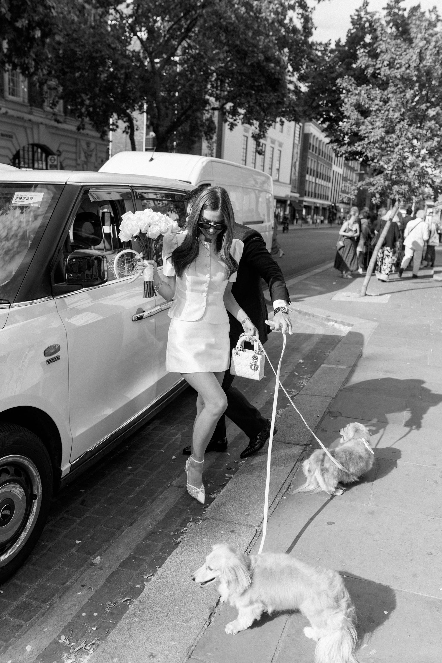 Bride in white mini dress walks two corgis beside a taxi on a London street.