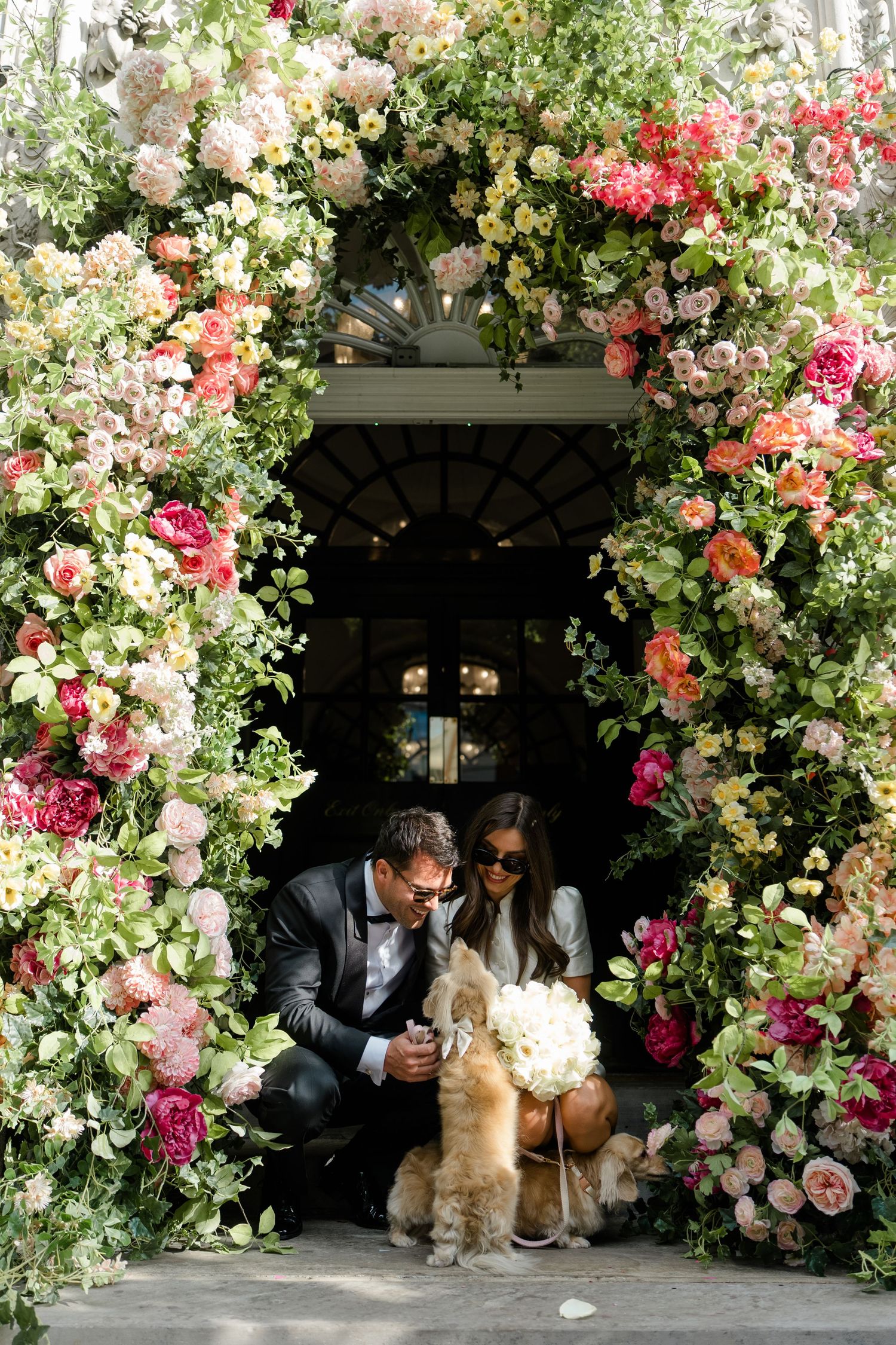 Bride and groom crouch with their dog beneath stunning floral arch of colorful roses at wedding venue.