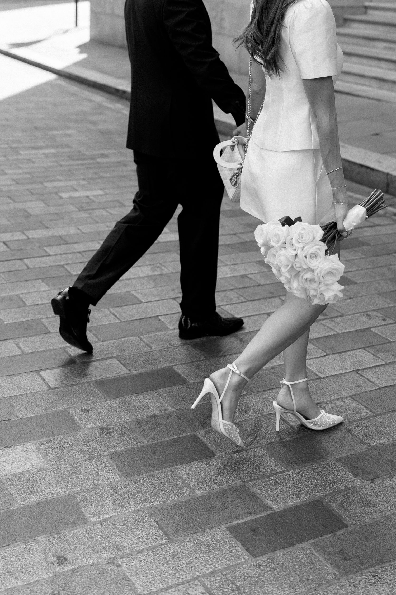 Black and white photo of bride and groom walking holding hands, bride carrying white bouquet.