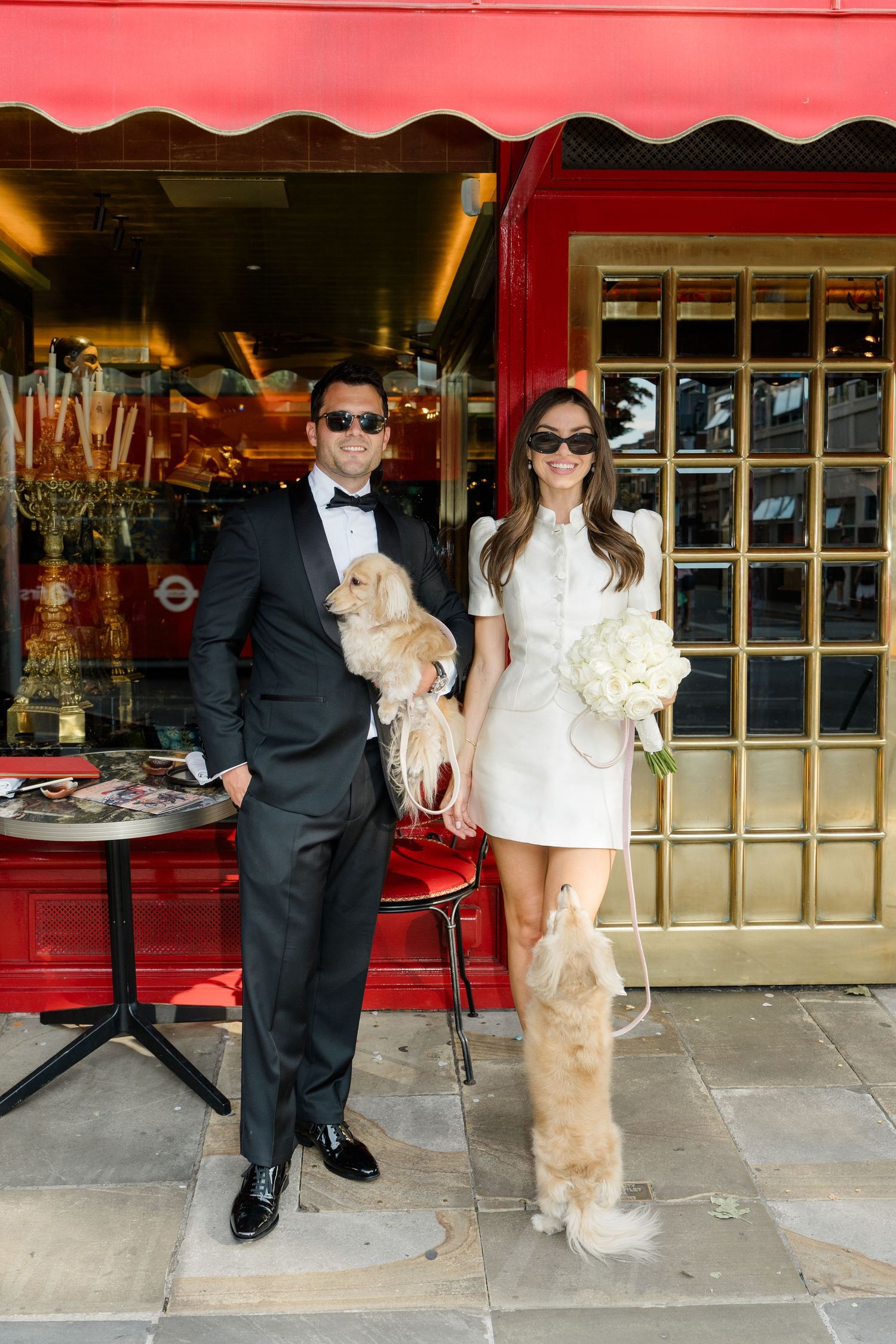 Bride and groom in sunglasses pose with fluffy dogs outside red-awning restaurant in London.