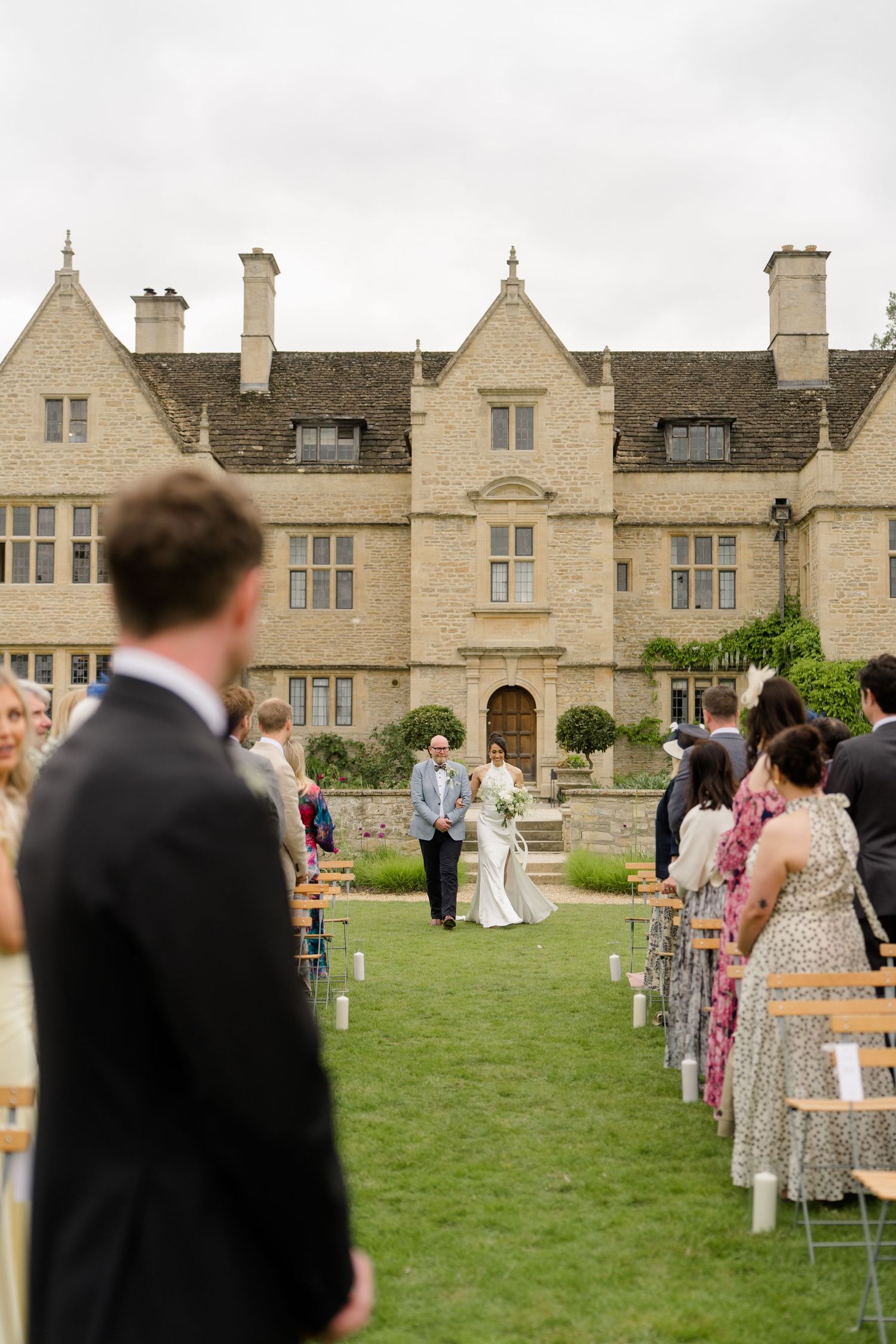 A bride walks down the outdoor aisle toward the groom at a grand English manor house wedding ceremony.