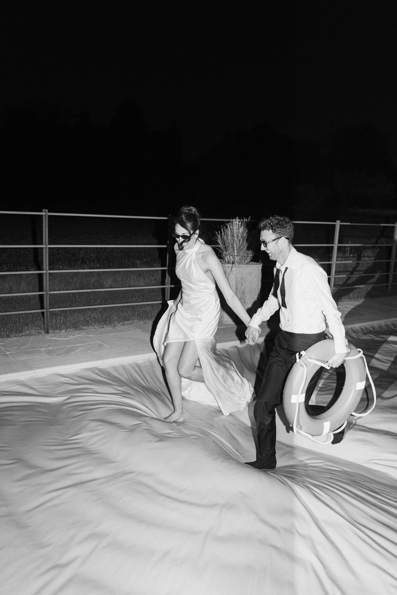 Black and white photo of bride and groom running across covered pool holding life preserver.