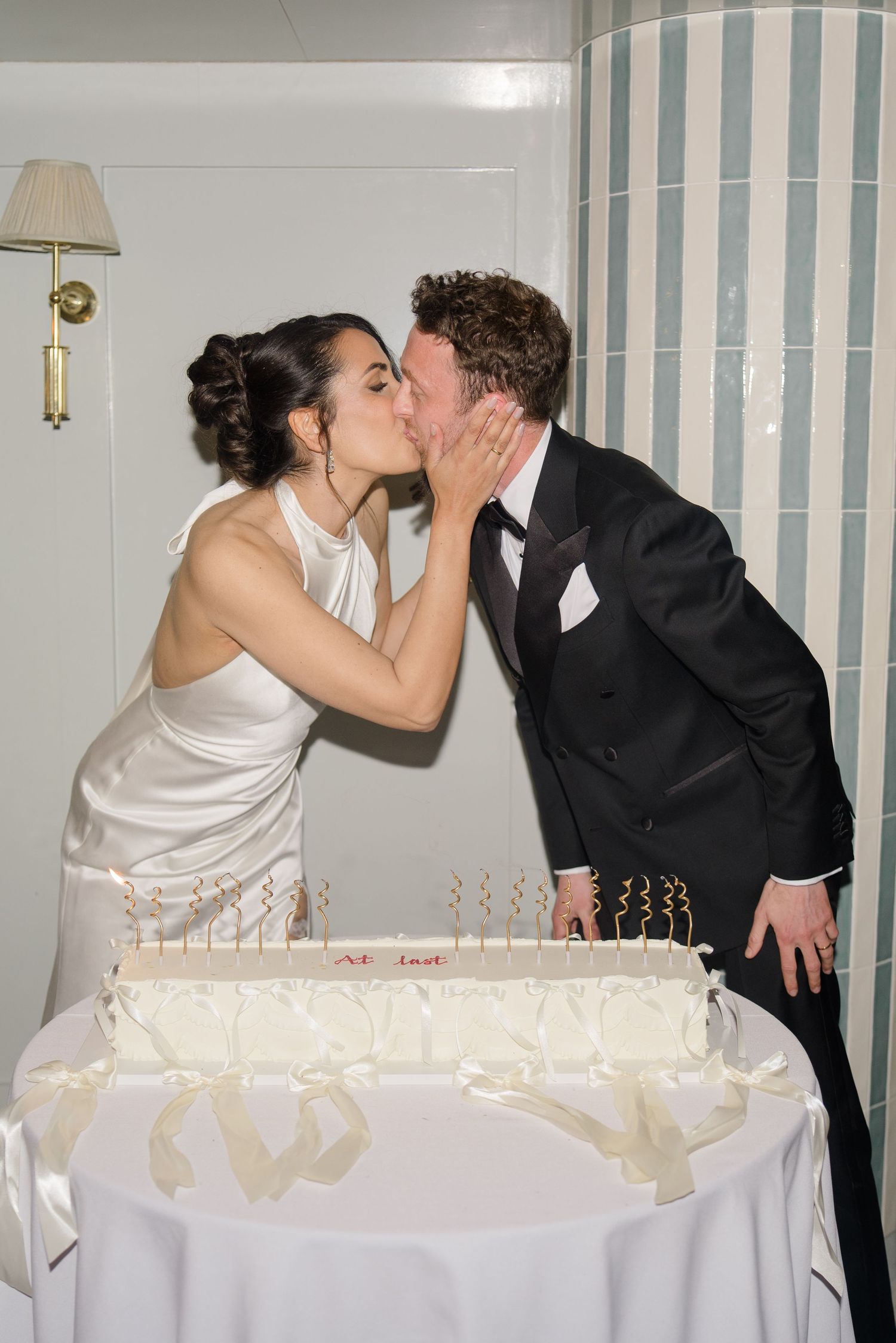 Bride and groom sharing a kiss over elegant white wedding cake with candles at reception.