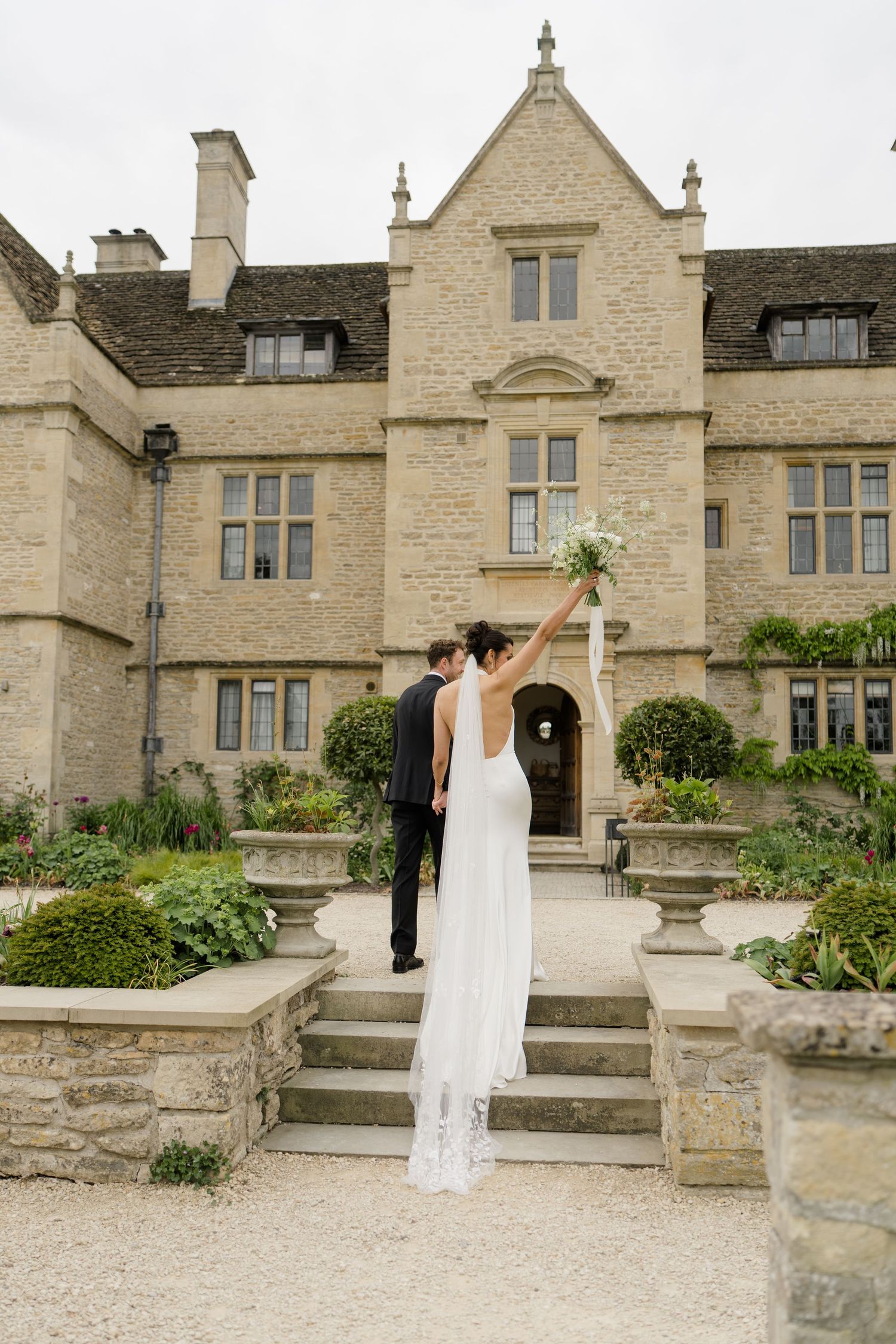 Bride raises bouquet triumphantly as couple stands before a grand Cotswolds stone manor house.