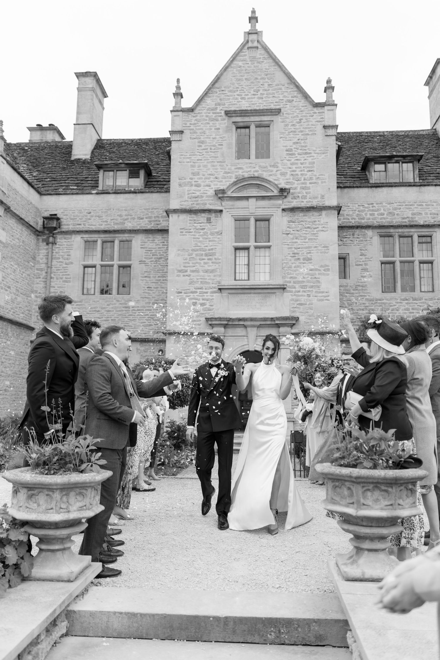 Bride and groom walking through a confetti shower outside a stunning historic stone manor house.