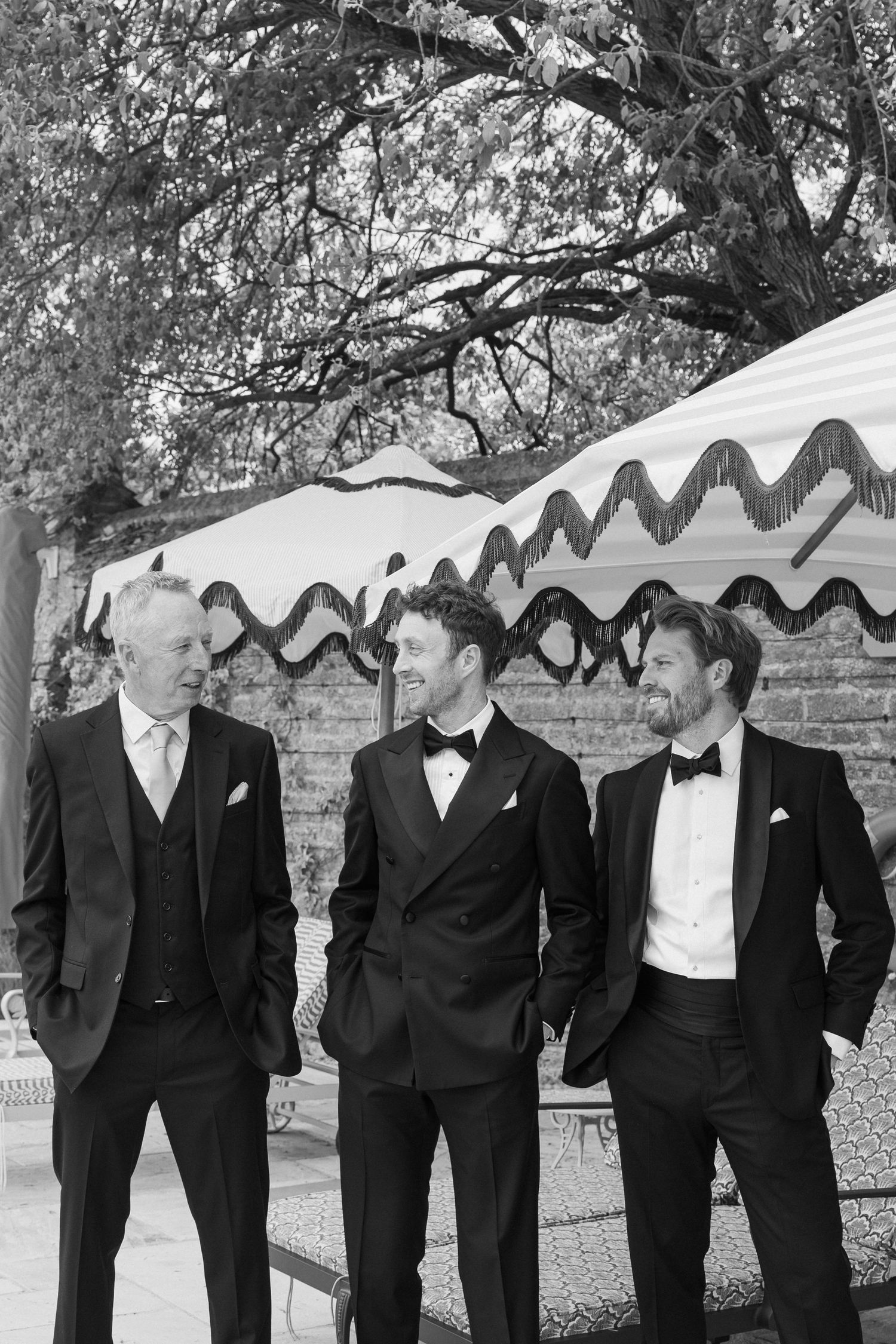 Three groomsmen in black tuxedos with bow ties laughing together outdoors at a wedding venue.