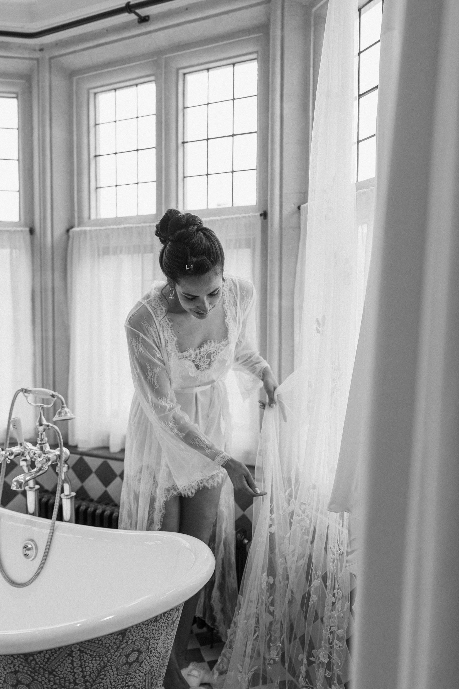 Bride in lace bridal robe adjusts her wedding dress beside a clawfoot bathtub in black and white photo.