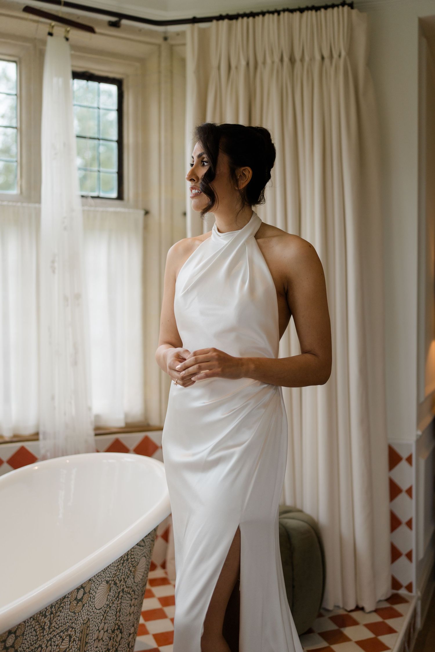 Bride in elegant white halter neck wedding dress standing by a clawfoot bathtub in a tiled bathroom.