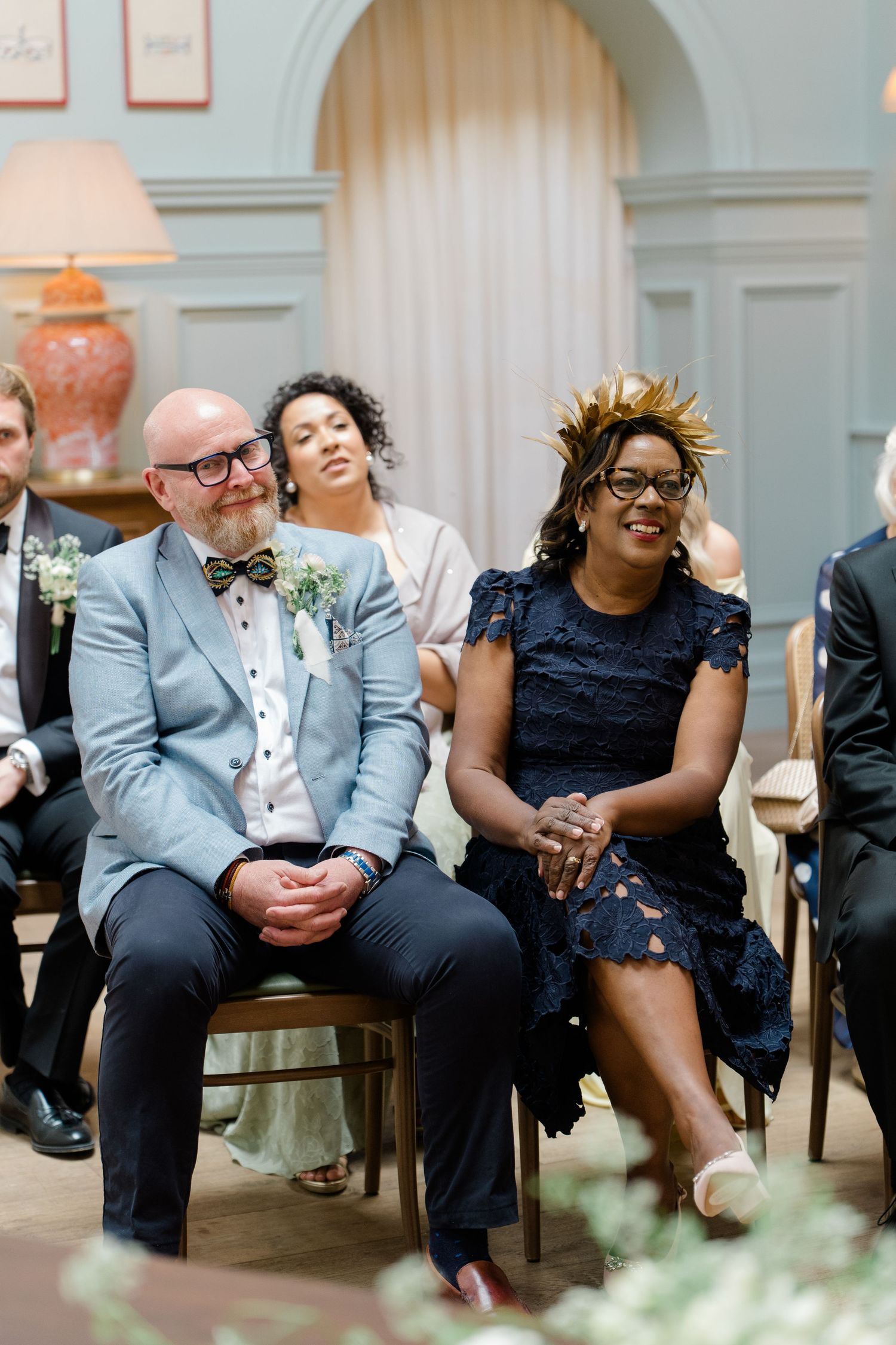 Wedding guests smile and laugh during a ceremony, woman in navy lace dress and man in gray suit with bow tie seated in chairs.