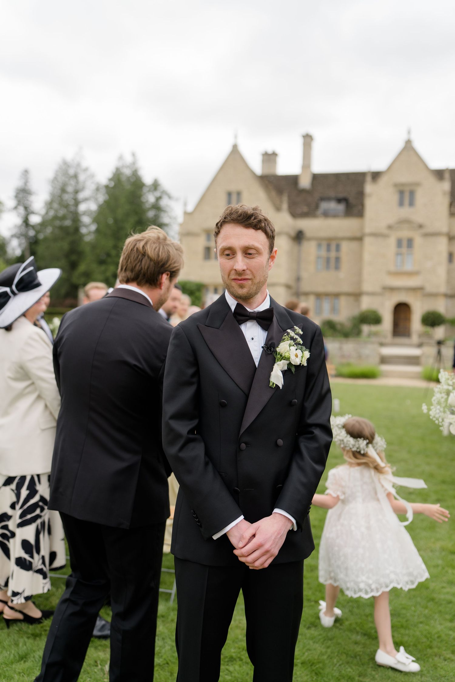 A groom in a black tuxedo with boutonniere stands outdoors at a wedding venue with guests and a flower girl nearby.