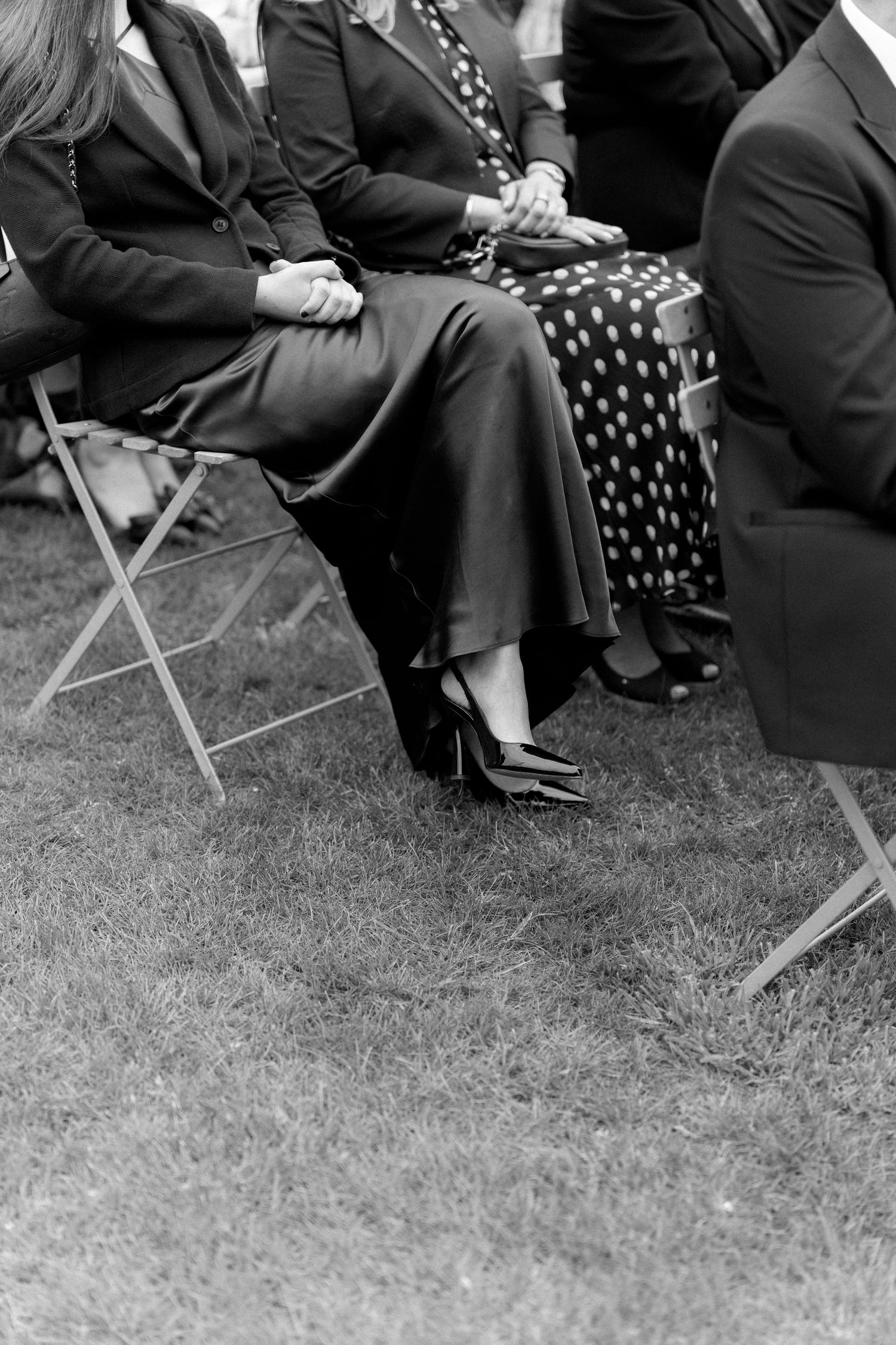 Black and white photo of elegantly dressed wedding guests seated on folding chairs on a grass lawn.