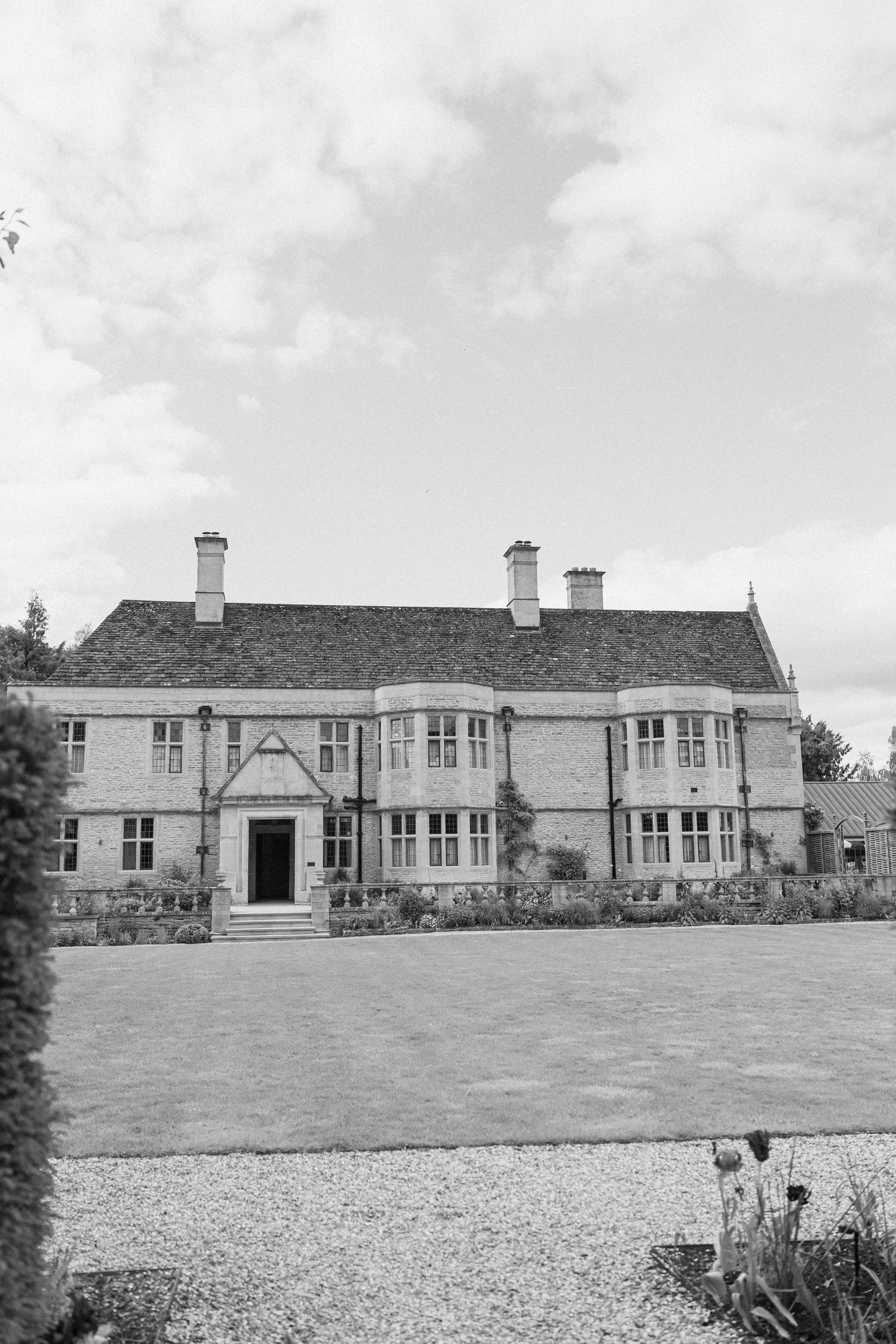 Black and white photo of a grand English country manor house with manicured lawn and gravel path.