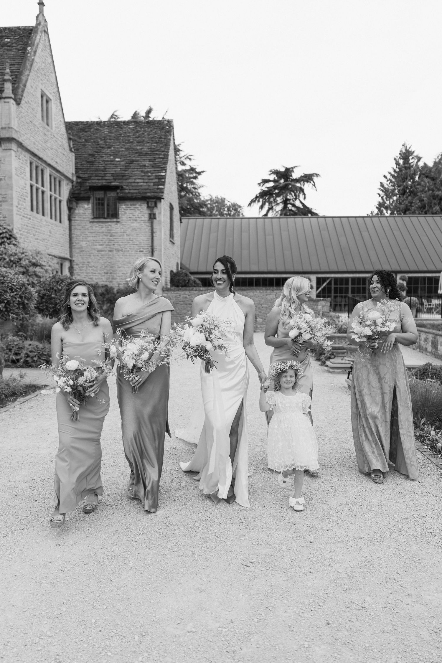 Bride walks with bridesmaids in sage gowns and flower girl outside historic English manor, black and white.