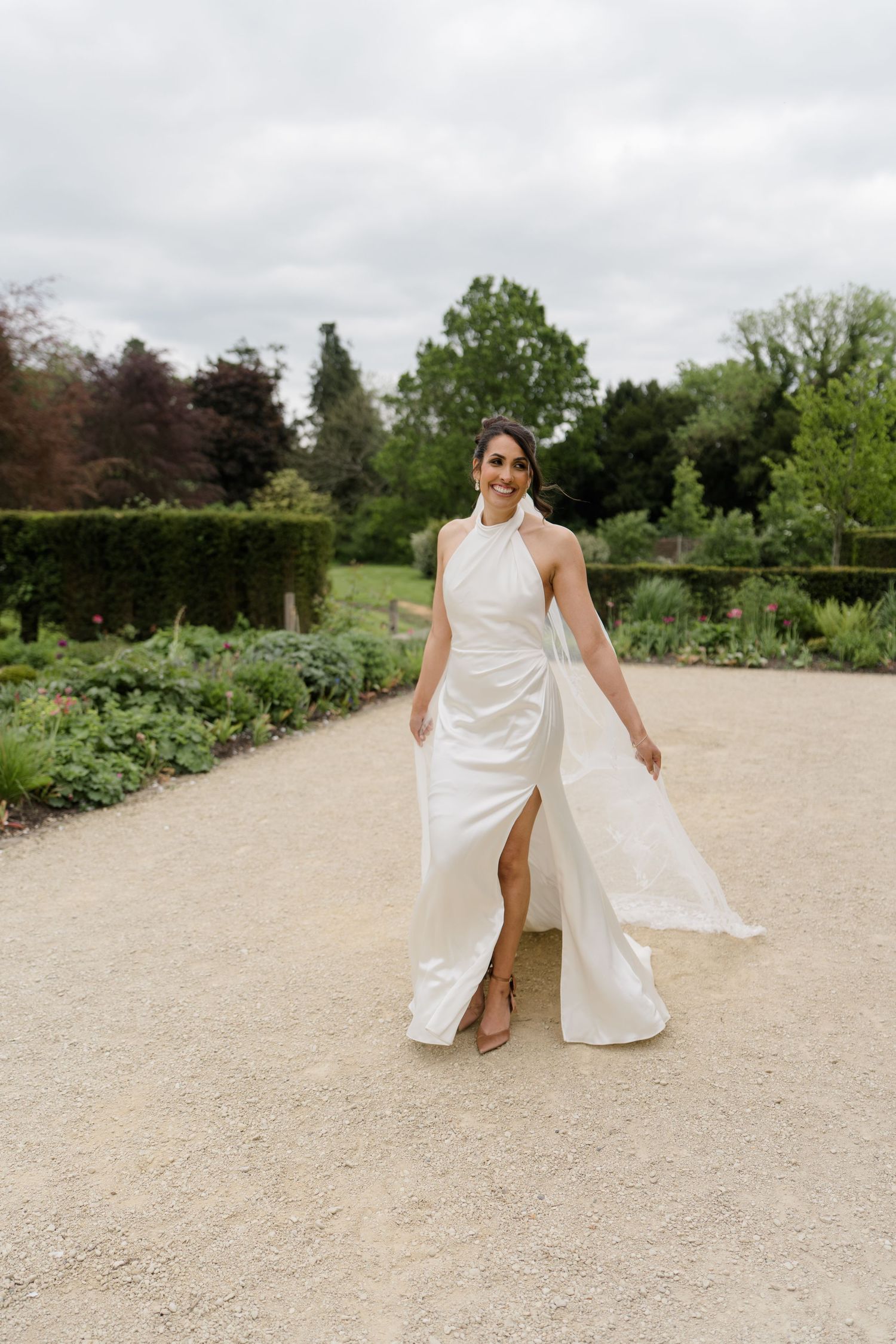 Bride in sleek white halter neck wedding dress with flowing fabric walks through formal garden path.