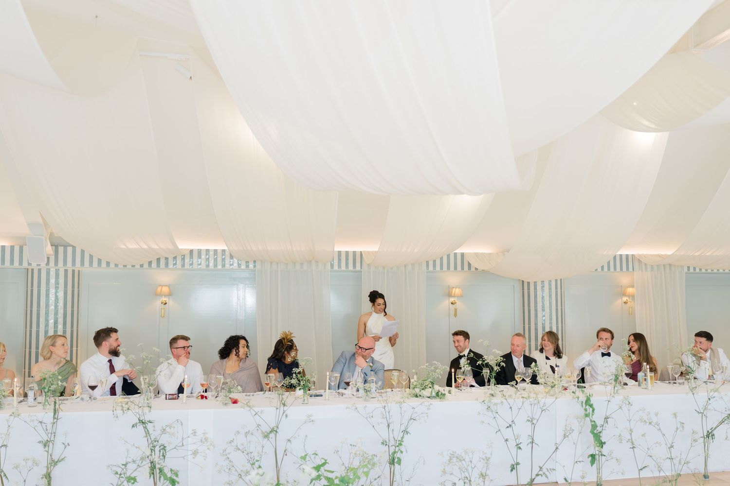 Bride gives speech at long head table during wedding reception in elegant draped marquee venue.
