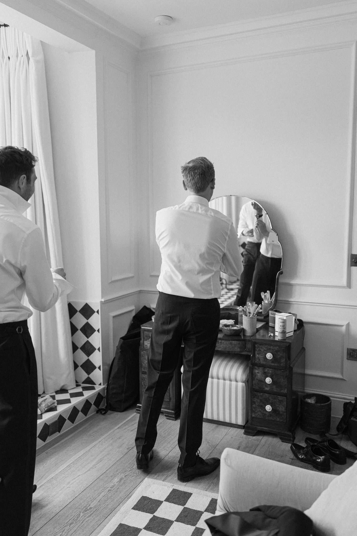Groom in white dress shirt checking his appearance in a vintage mirror atop a dark wooden dresser.