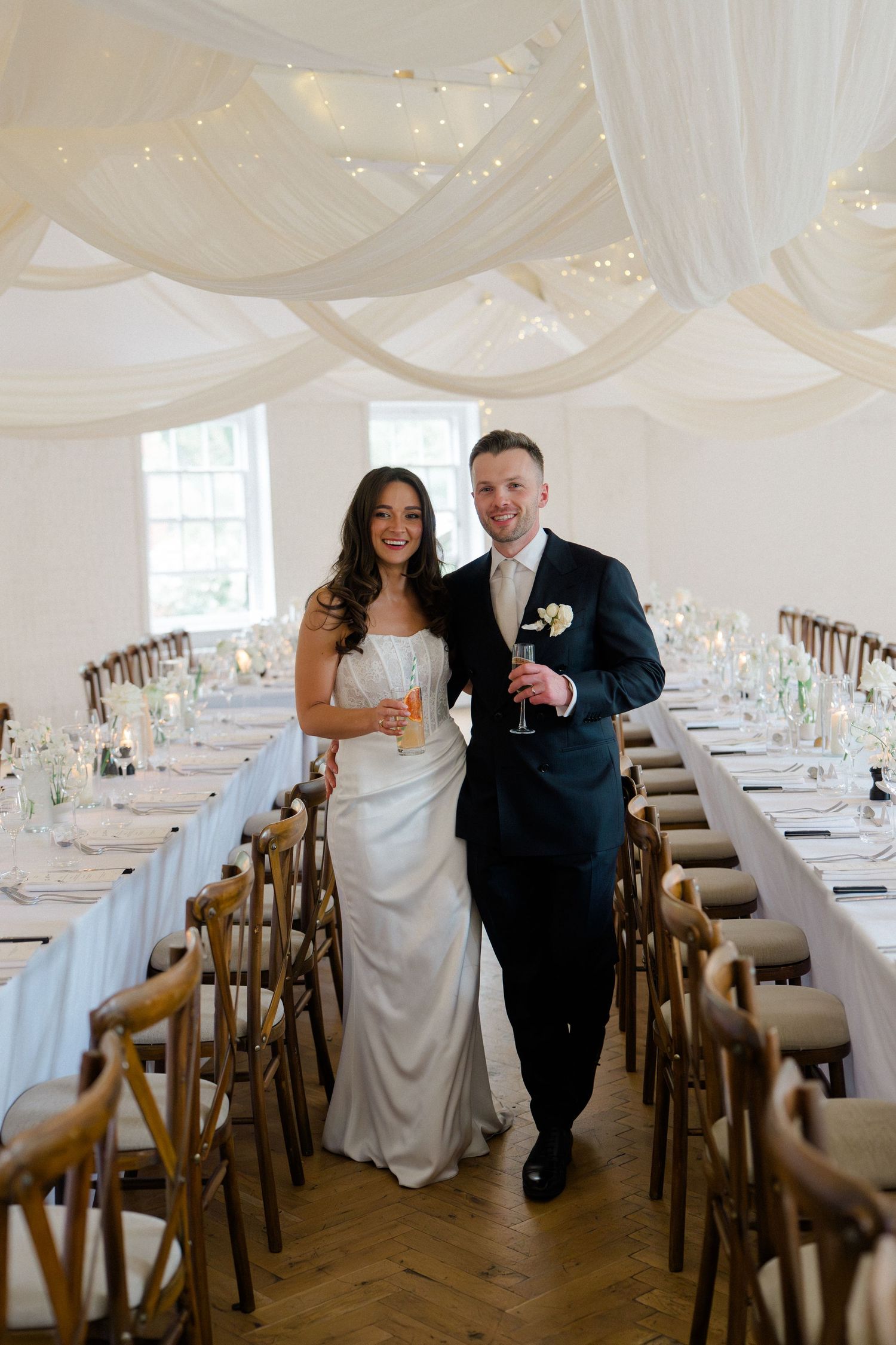 Bride and groom smiling in elegant wedding reception hall with white draped ceiling and fairy lights.