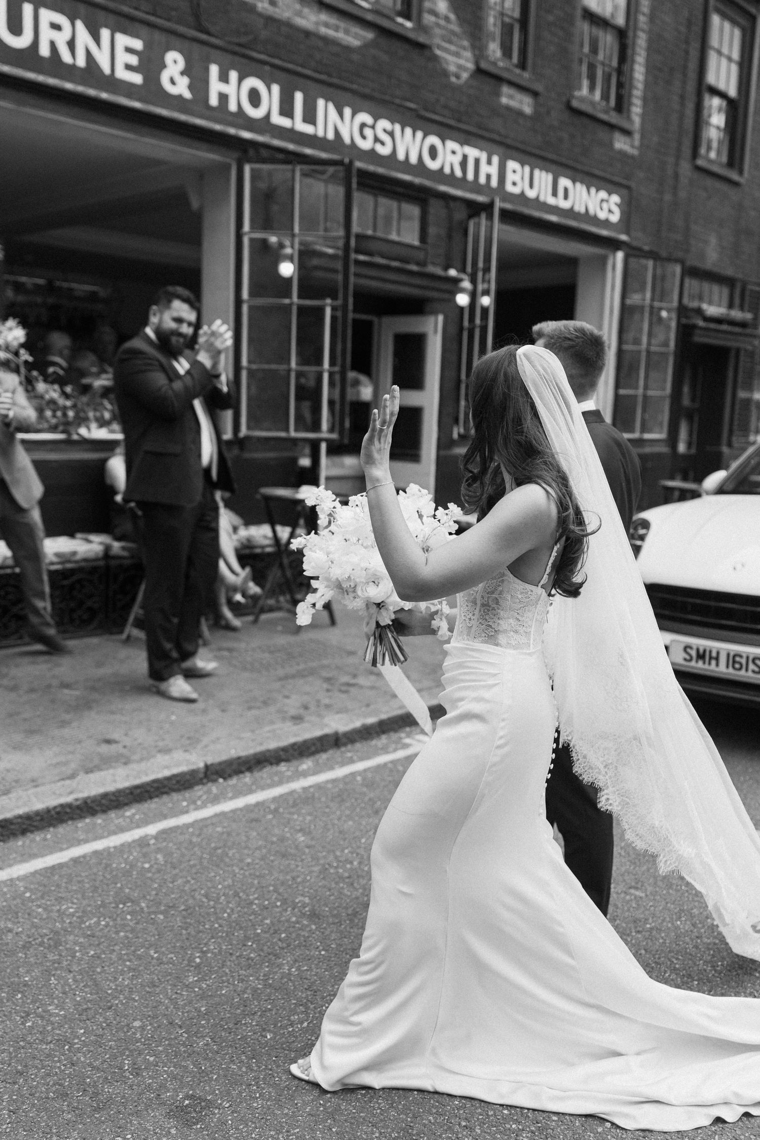 Bride in elegant white gown and veil waves outside Bourne and Hollingsworth Buildings.