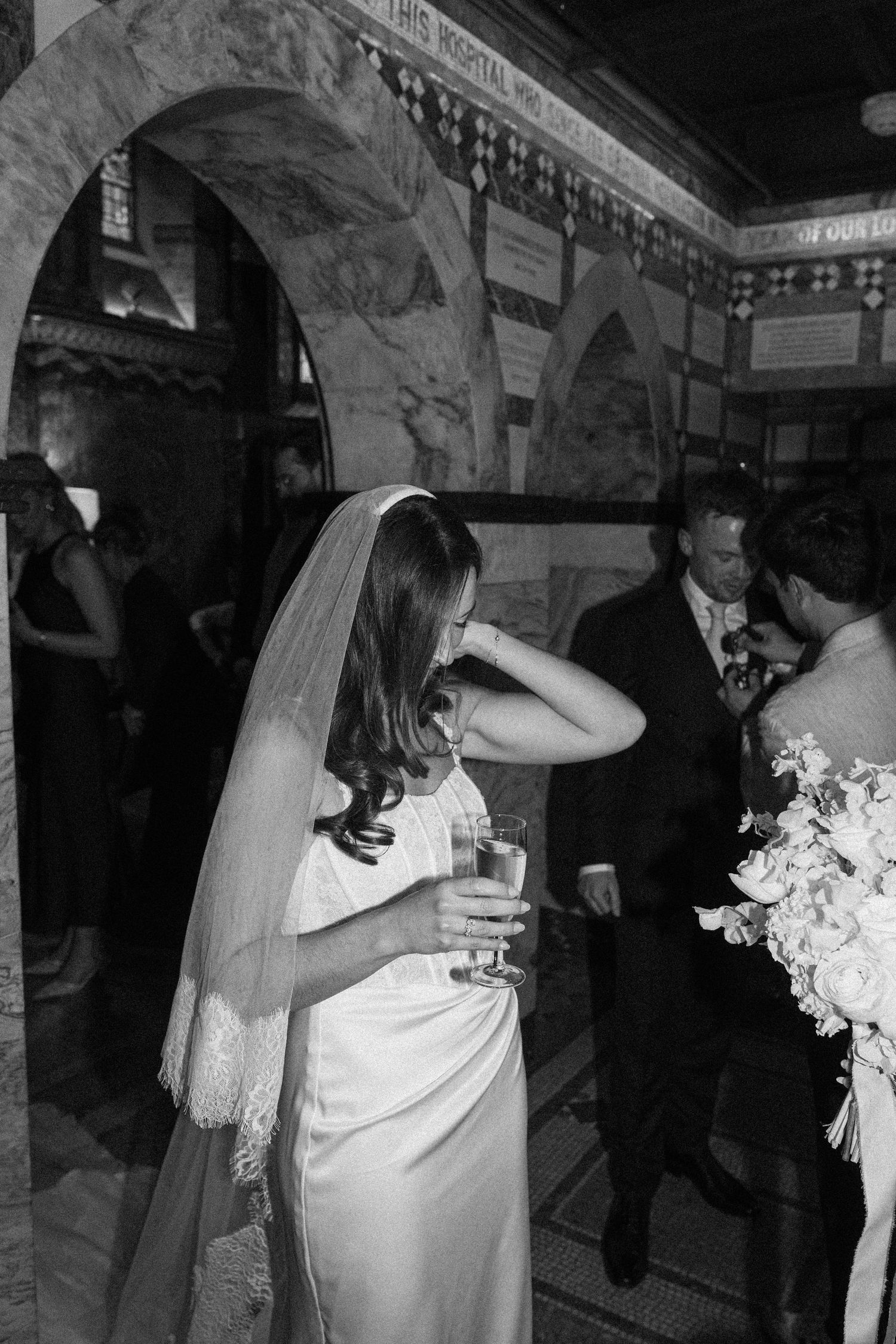 Bride in white gown and veil holding champagne glass under stone arch at wedding reception.