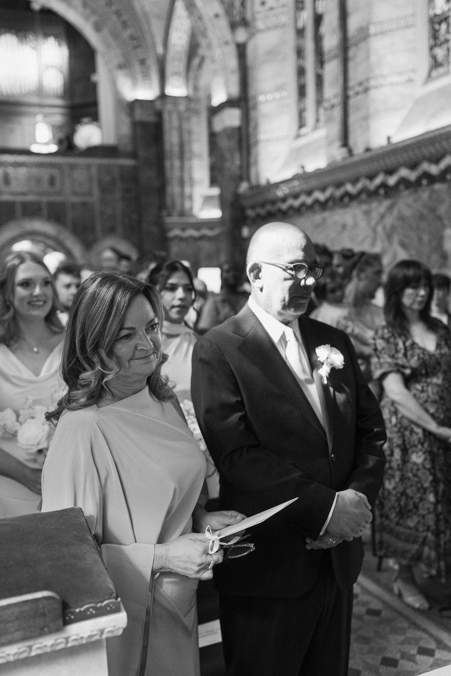 Black and white photo of wedding guests watching ceremony inside ornate church, man in suit with boutonniere.